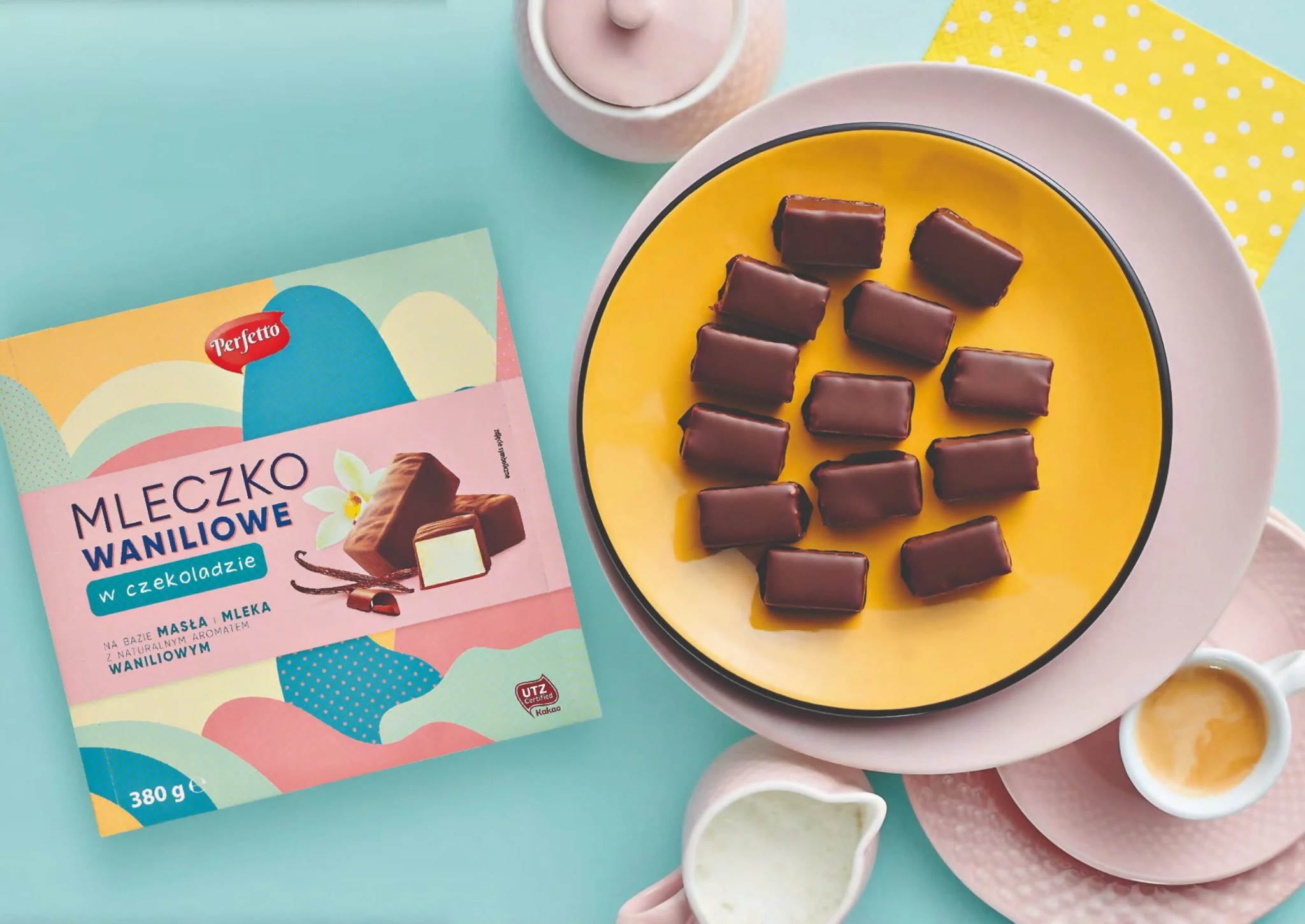 Stylizowana kompozycja cukierków Mleczko Waniliowe Perfetto na żółtym talerzu, obok opakowanie produktu, filiżanka z kawą i dzbanek z mlekiem, na tle w pastelowych kolorach.