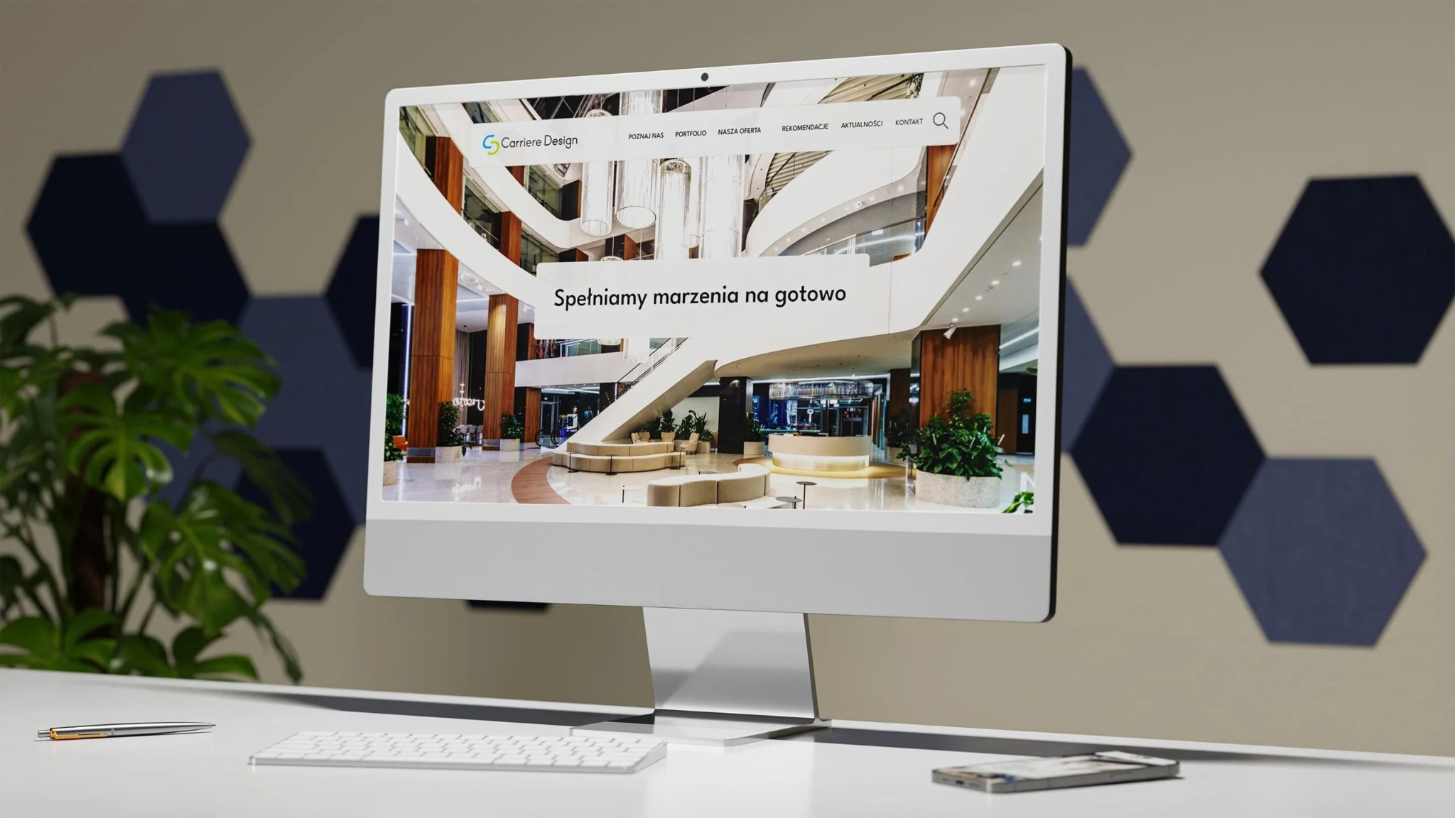 Ekran komputera iMac prezentuje stronę internetową Carriere Design z widokiem na eleganckie wnętrze hotelu lub biurowca; roślina doniczkowa, klawiatura i długopis na białym biurku...