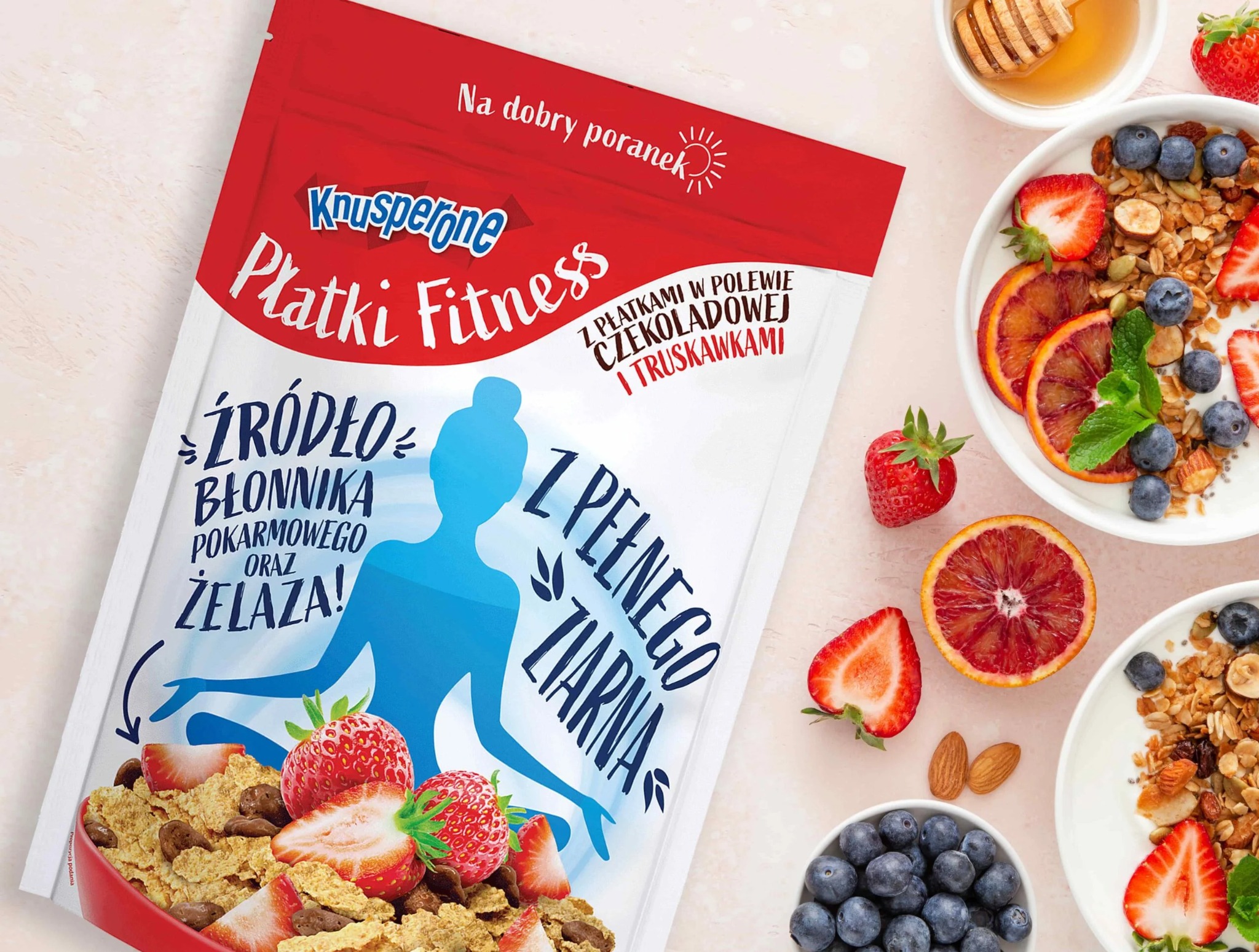 Opakowanie płatków śniadaniowych Knusperone Fitness z widocznym napisem 'Źródło błonnika pokarmowego oraz żelaza!', obok miseczki z jogurtem, granolą, krwistymi pomarańczami, borówkami...