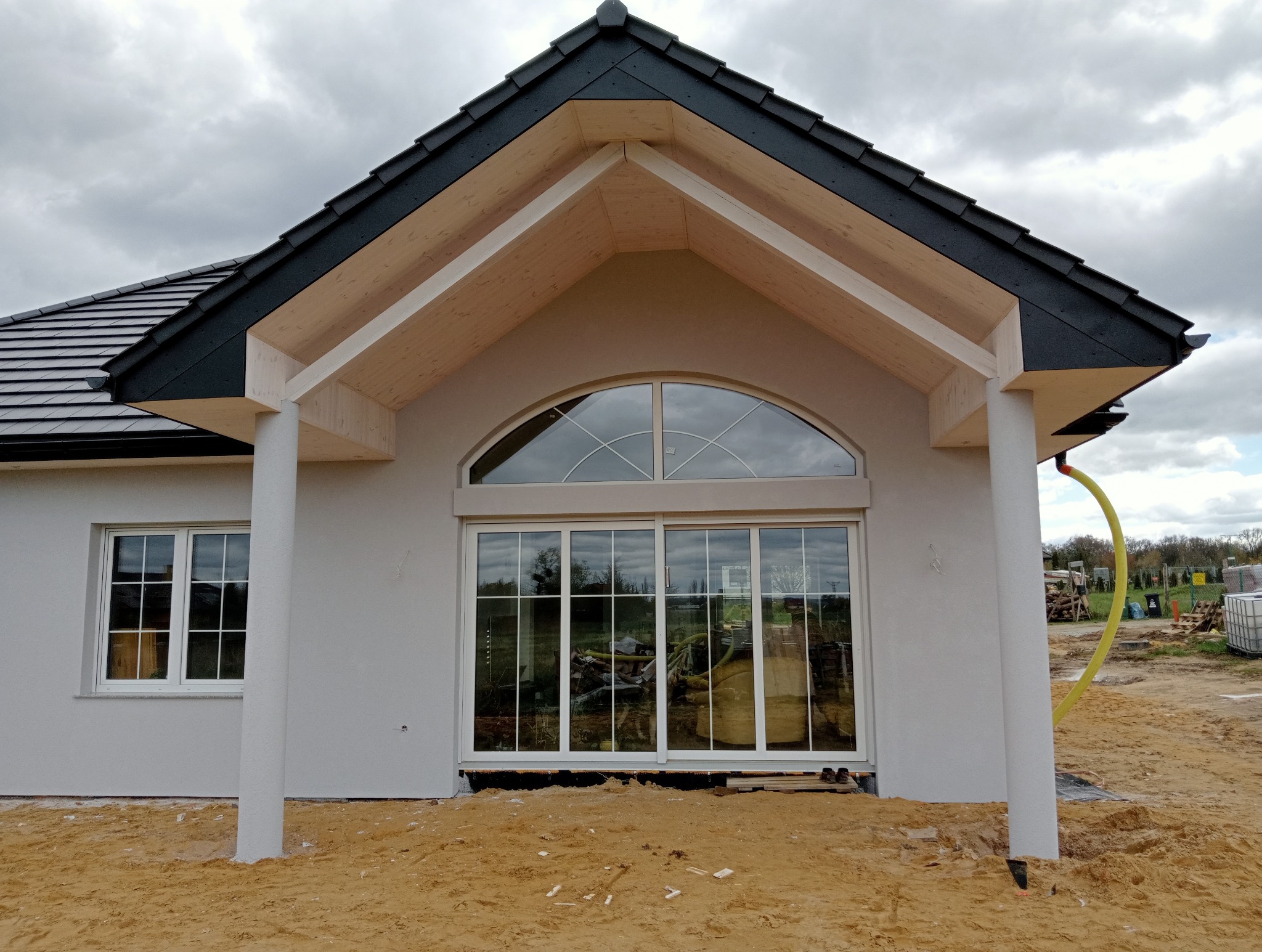 Fragment elewacji domu jednorodzinnego z dużym oknem łukowym i kolumnami, świeżo otynkowany, z widocznymi śladami prac budowlanych wokół.