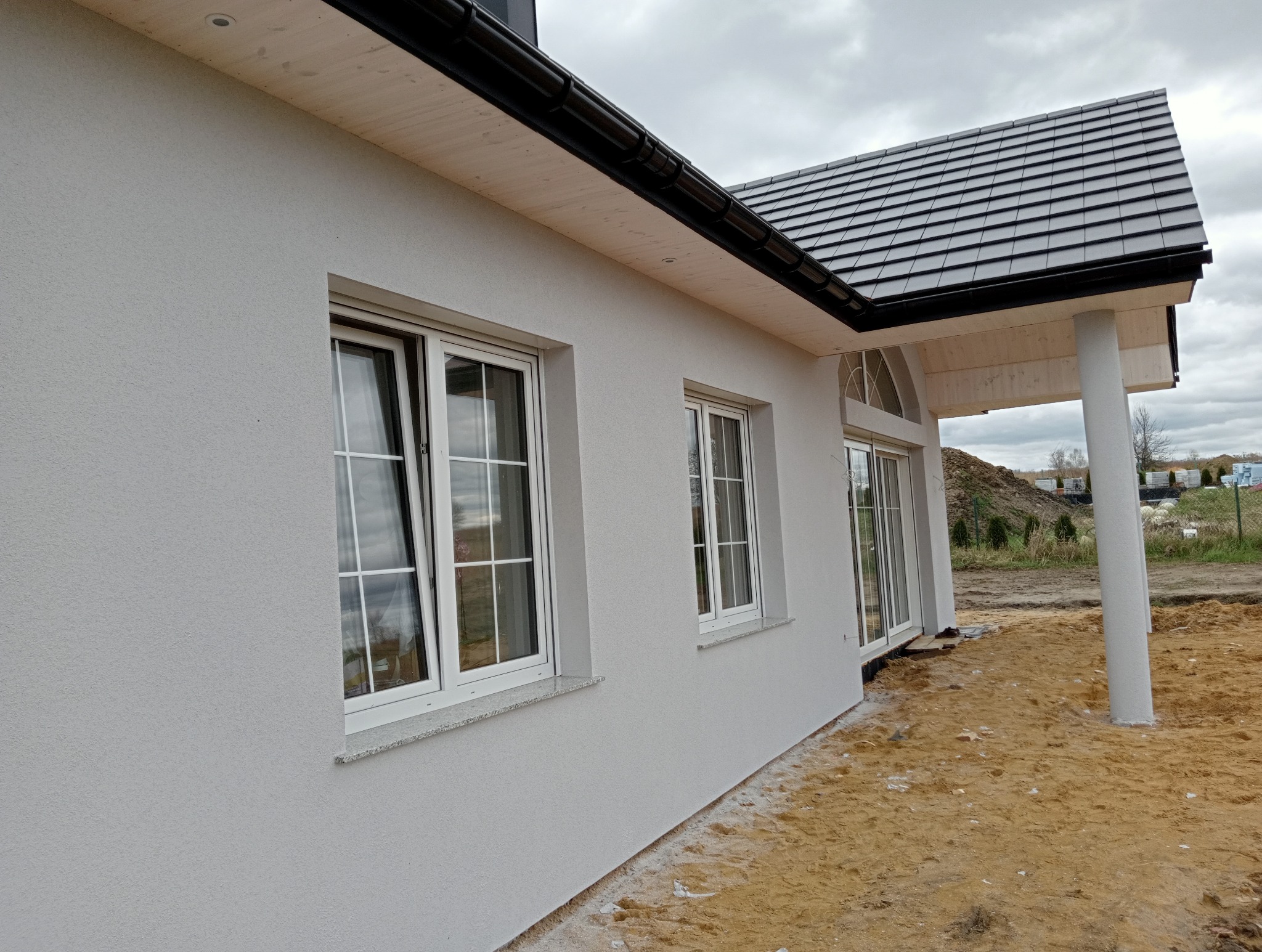 Jasnoszara elewacja domu jednorodzinnego z białymi oknami i ciemną dachówką, widok z boku na plac budowy z piaskiem.