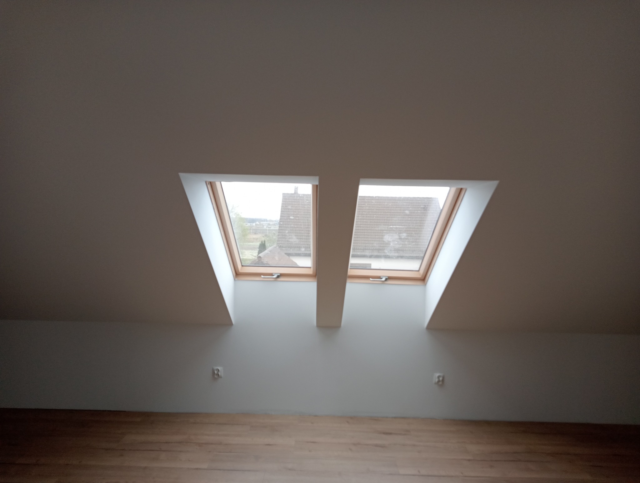 Poddasze z dwoma oknami dachowymi, drewniana podłoga, białe ściany i skosy sufitu, widok na zewnątrz przez okna.