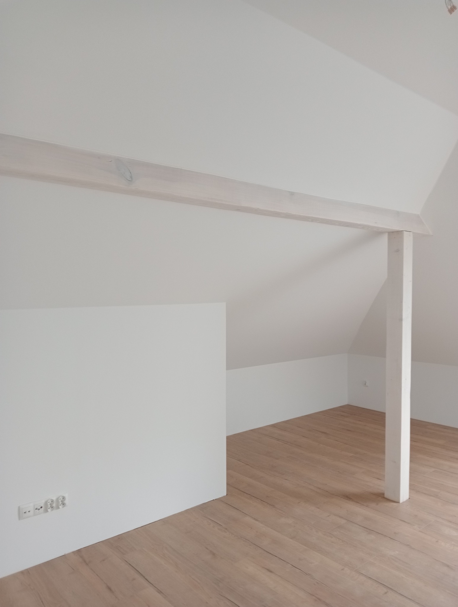 Poddasze po remoncie: drewniana podłoga, białe ściany, pomalowana belka konstrukcyjna i słup, gniazdka elektryczne przy podłodze.