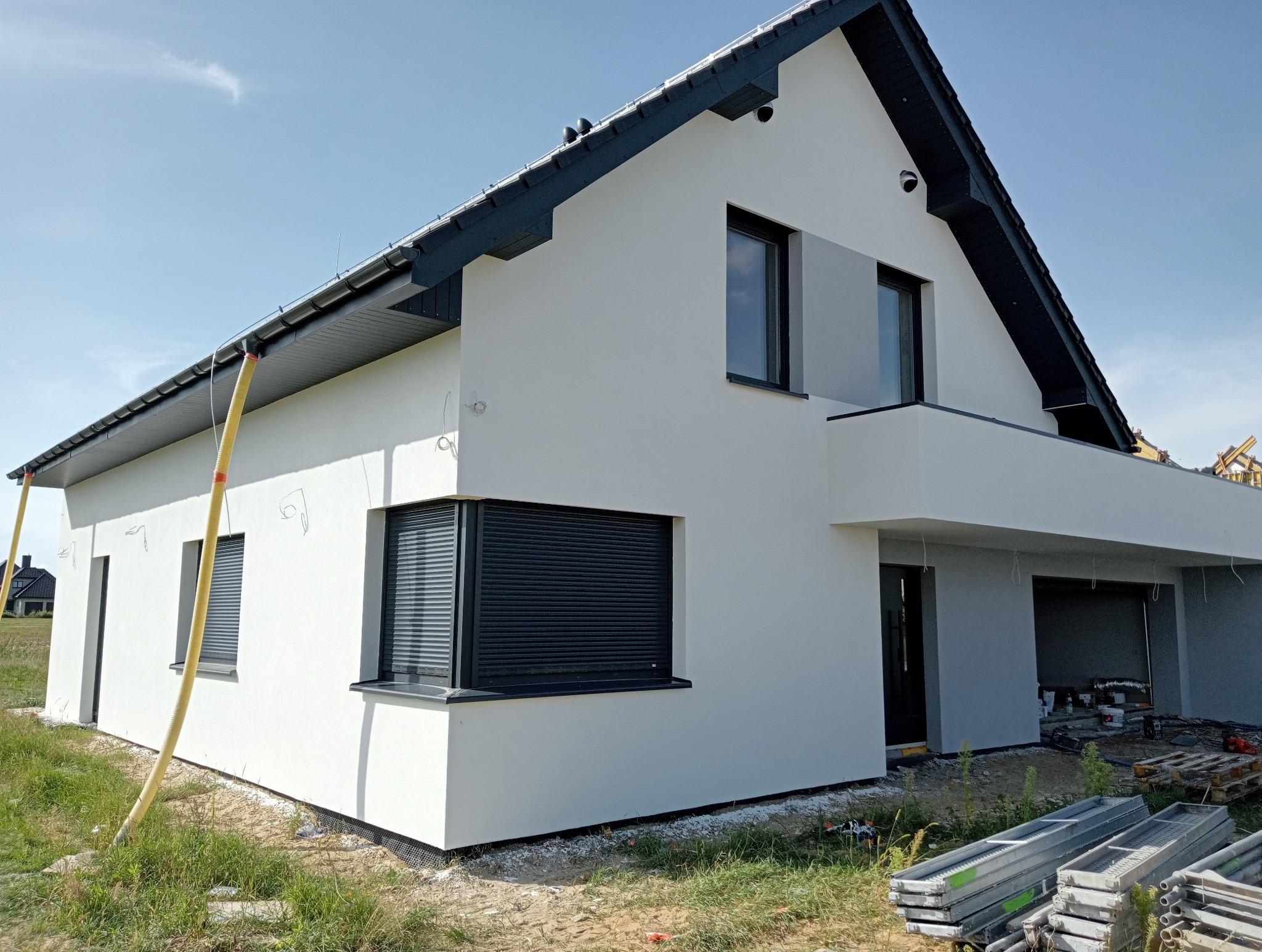 Nowoczesny dom jednorodzinny z białą elewacją, czarnymi ramami okiennymi i roletami zewnętrznymi, widoczne przewody wentylacyjne do osuszania ścian po pracach wykończeniowych, teren wokół domu...