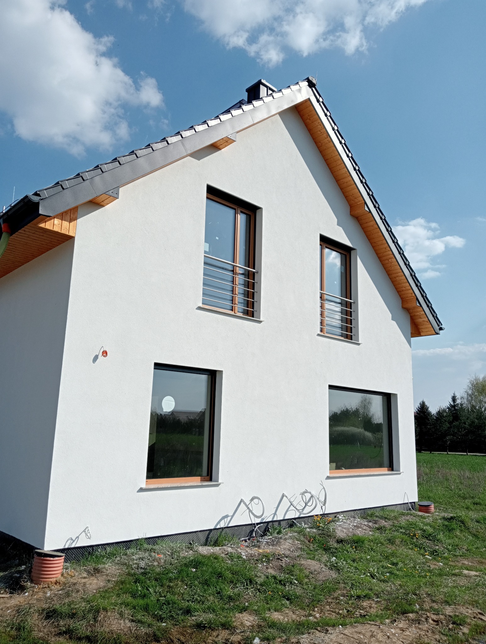 Nowoczesny dom jednorodzinny z białym tynkiem zewnętrznym, widok z boku, z widocznymi oknami z drewnianymi ramami i metalowymi balustradami, szara dachówka, elementy instalacji wystające z ziemi.