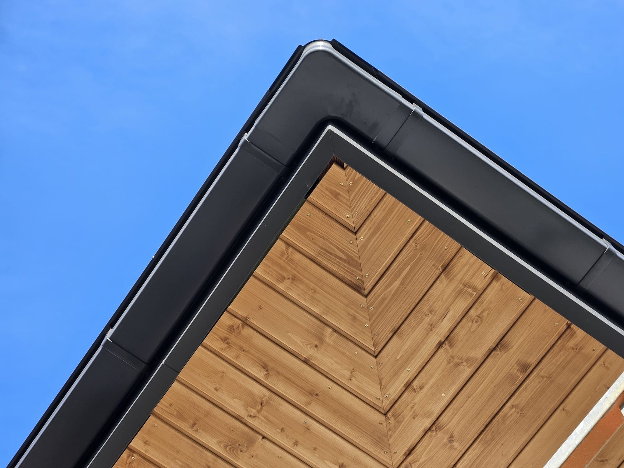 Szczegółowe ujęcie rogu budynku z drewnianą elewacją ułożoną w jodełkę i czarną, metalową rynną na tle błękitnego nieba.