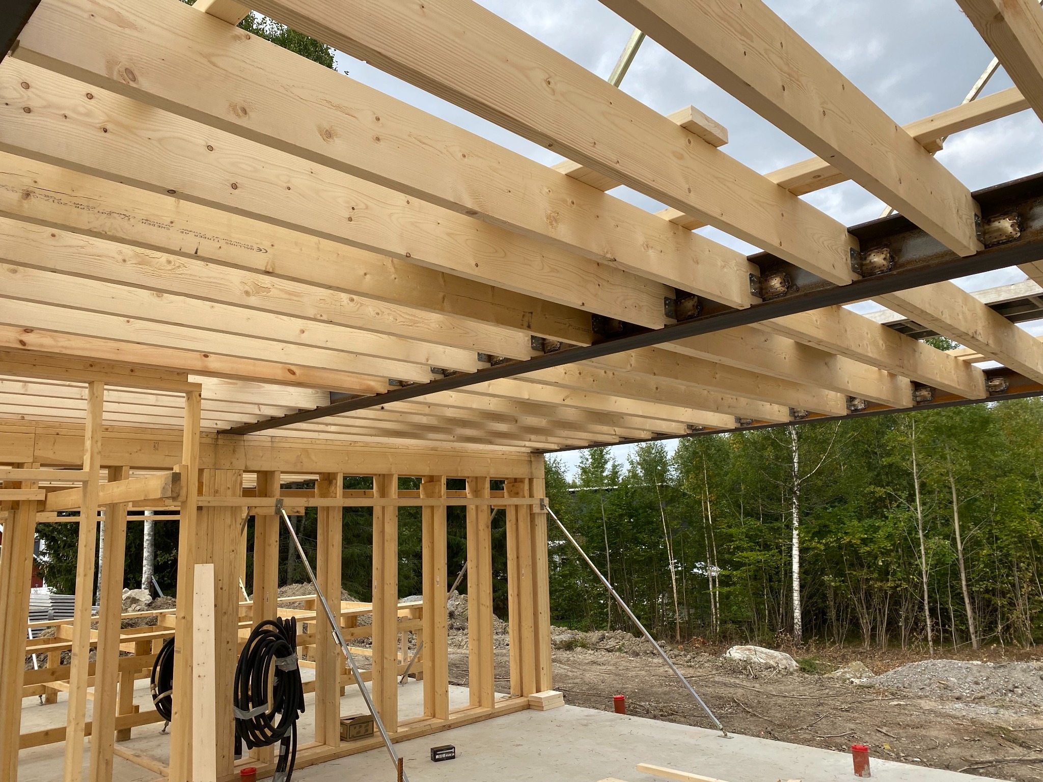 Wnętrze domu w trakcie budowy, widoczne drewniane belki stropowe mocowane do stalowej konstrukcji, ściany szkieletowe oraz zwisające kable elektryczne.