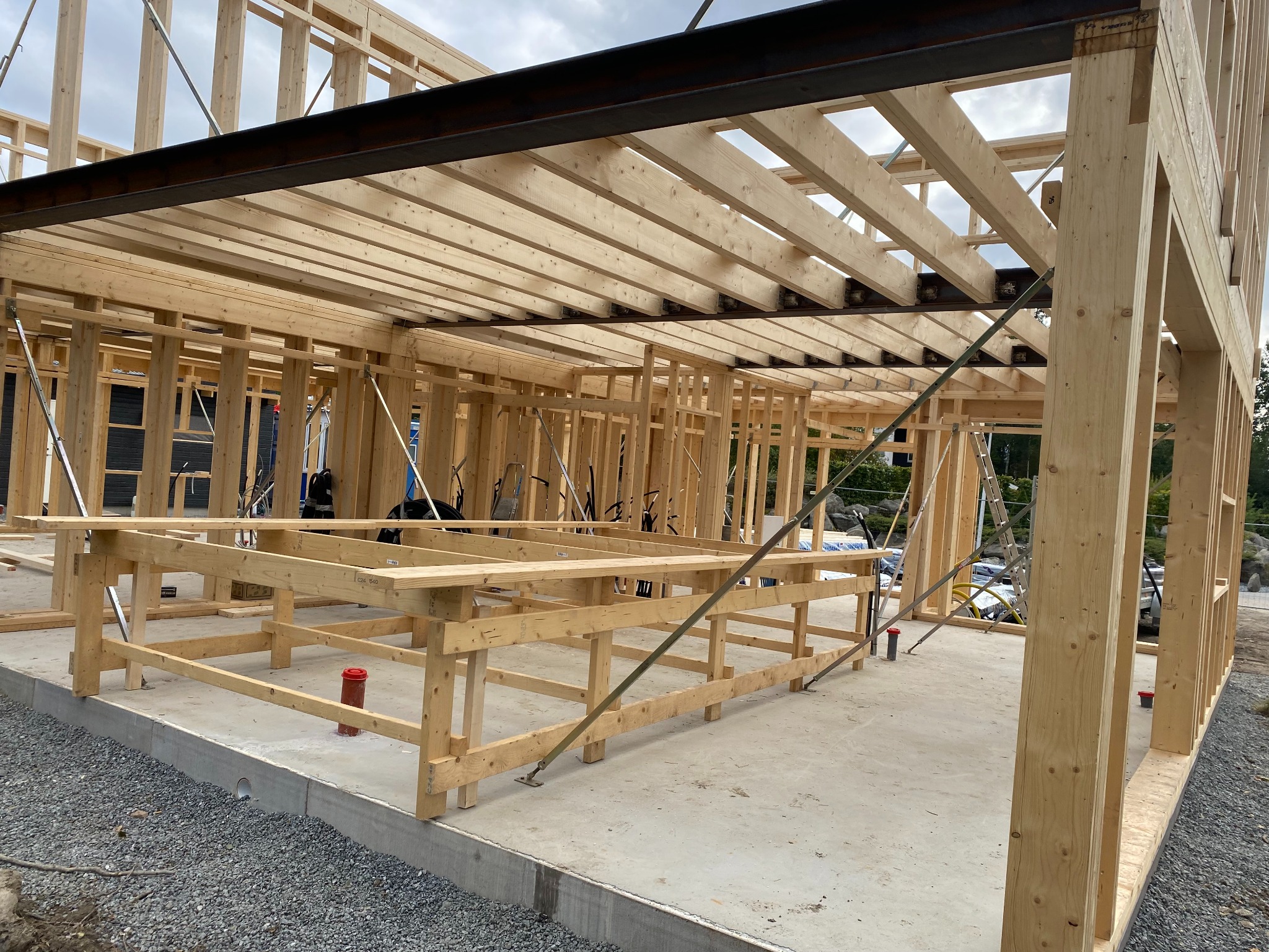 Wnętrze domu w trakcie budowy w technologii szkieletowej, widoczne drewniane ramy ścian i stropu połączone z elementami stalowymi, betonowa podłoga z widocznymi instalacjami.