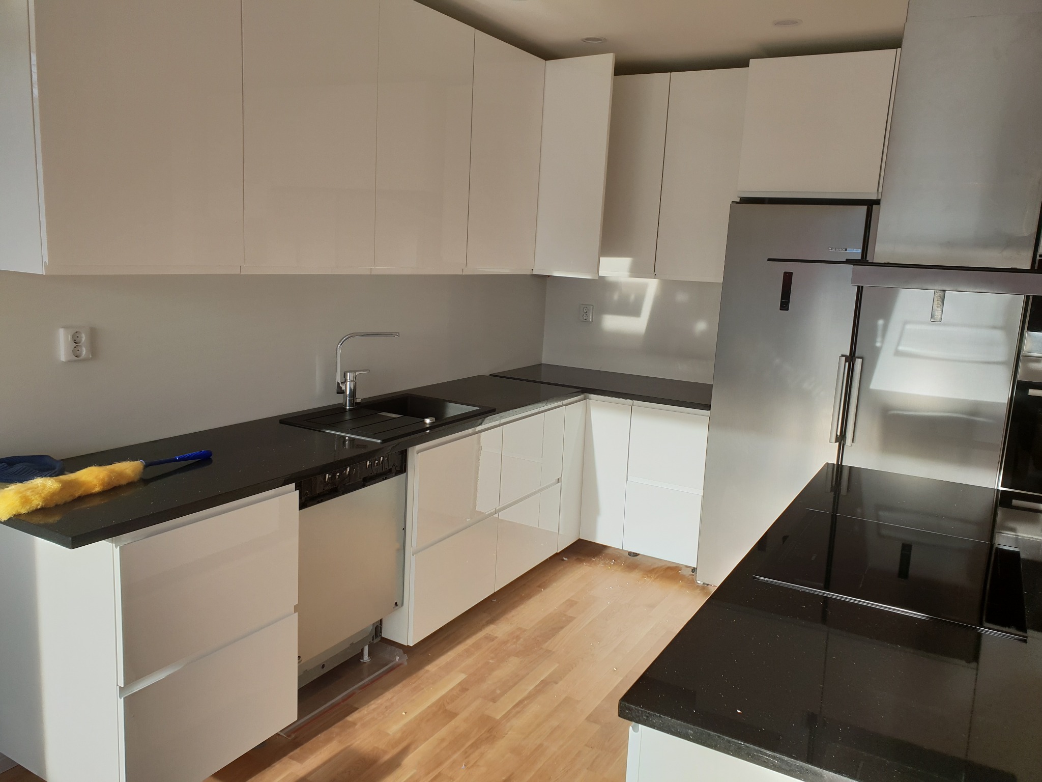 Jasna kuchnia z białymi, błyszczącymi szafkami i czarnym blatem, widoczny fragment podłogi z jasnego drewna, na blacie leży żółta miotełka do kurzu. W tle srebrna lodówka i okap.