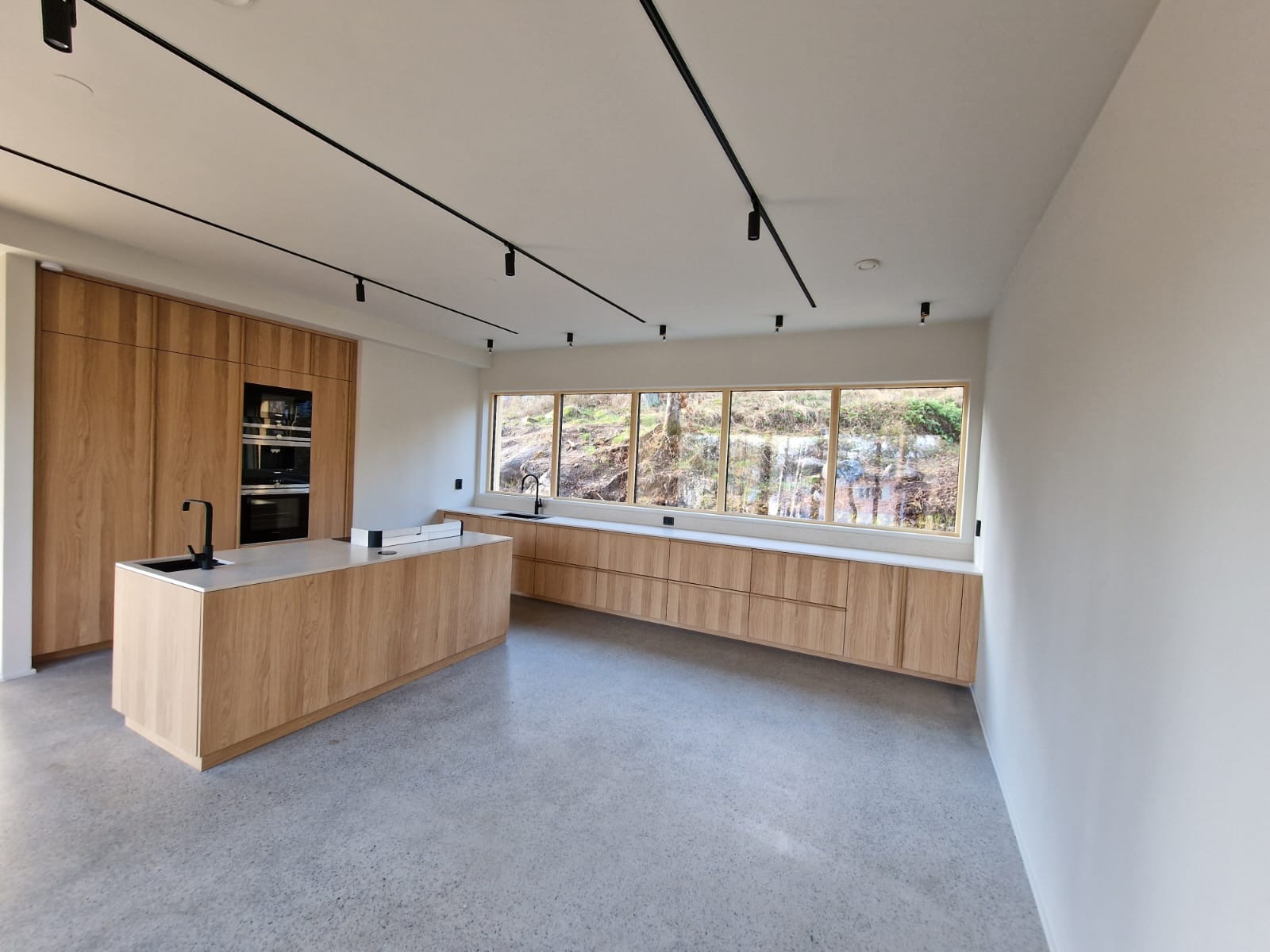 Nowoczesna kuchnia z drewnianymi szafkami, wyspą i dużym oknem z widokiem na zalesione wzgórze. Czarne szyny oświetleniowe na suficie.
