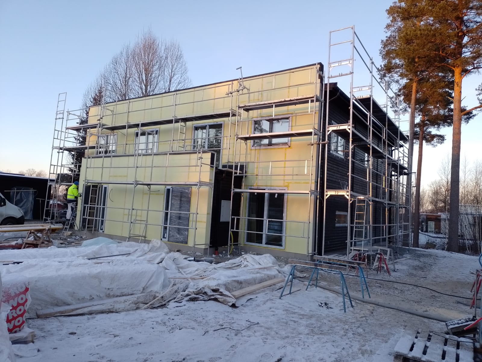 Dwukondygnacyjny dom w trakcie budowy, z ociepleniem zewnętrznym i rusztowaniami, na zimowym placu budowy.