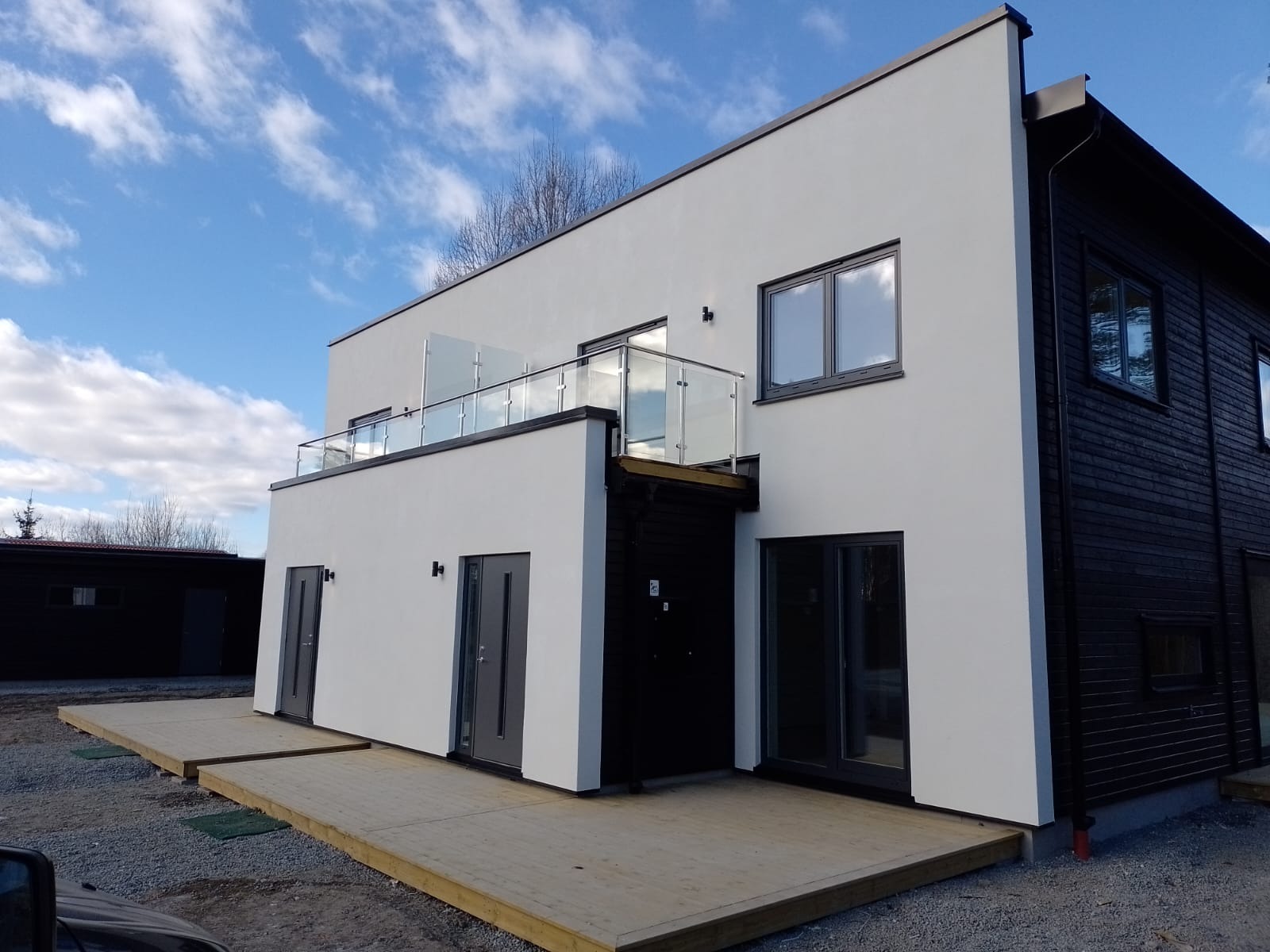 Nowoczesny dwukondygnacyjny dom z białym tynkiem i czarną drewnianą elewacją, balkon z przeszkloną balustradą, drewniany taras na poziomie gruntu.