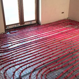 Piotr Groński AIRCOOL TECH - Czerwone rury ogrzewania podłogowego ułożone na folii z nadrukiem kratki, widoczne okno i fragment poddasza. Instalacja przed wylaniem posadzki.