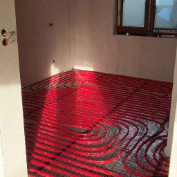 Piotr Groński AIRCOOL TECH - Czerwone rury ogrzewania podłogowego marki Warmit ułożone na folii w jasnym pomieszczeniu z oknem. Widoczne elementy instalacji elektrycznej.
