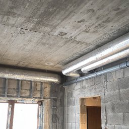 Piotr Groński AIRCOOL TECH - Surowy strop z widocznymi rurami wentylacyjnymi w srebrnej izolacji i szarymi rurami PCV, zamontowanymi w surowym wnętrzu budynku z szarej cegły.