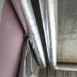 Piotr Groński AIRCOOL TECH - Dwa izolowane przewody wentylacyjne z folią aluminiową przymocowane do sufitu w surowym wnętrzu. Widoczne elementy konstrukcyjne ścian i okno.