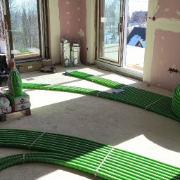 Piotr Groński AIRCOOL TECH - Wnętrze w trakcie prac instalacyjnych: zielone rury wentylacyjne ułożone na podłodze, materiały budowlane i narzędzia w tle, okna z widokiem na zewnątrz.