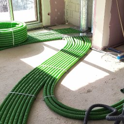 Piotr Groński AIRCOOL TECH - Zielone rury ogrzewania podłogowego ułożone w surowym wnętrzu, przygotowane do zalania posadzką. Widoczne okno i fragmenty ścian w trakcie wykończenia.