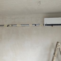 Piotr Groński AIRCOOL TECH - Montaż klimatyzacji w surowym wnętrzu: biała jednostka wewnętrzna Vivax zamontowana na ścianie, widoczne przewody w listwie i drabina malarska. Ściany w trakcie wykończenia.