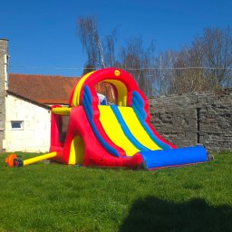 EventMagic - Rozłożona dmuchana zjeżdżalnia w kolorach czerwonym, żółtym i niebieskim na trawie, z podłączoną pompką elektryczną, na tle muru z kamienia i budynku z czerwoną dachówką.
