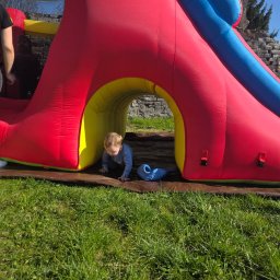 EventMagic - Chłopiec raczkuje w tunelu dmuchanego zamku z czerwoną ścianą, żółtym tunelem i niebieską zjeżdżalnią, na trawie w słoneczny dzień.