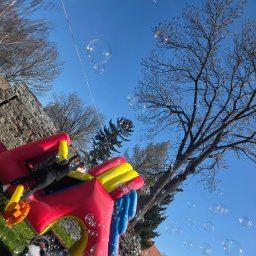 EventMagic - Dziewczynka w czarnym ubraniu leży na dmuchanym zamku w słoneczny dzień, w tle drzewa i budynki, a w powietrzu unoszą się bańki mydlane.