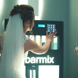 EventMagic - Panna młoda w białej sukni obsługuje automat do drinków Barmix, obok stoi mężczyzna z drinkiem w ręku, w tle butelki alkoholu.