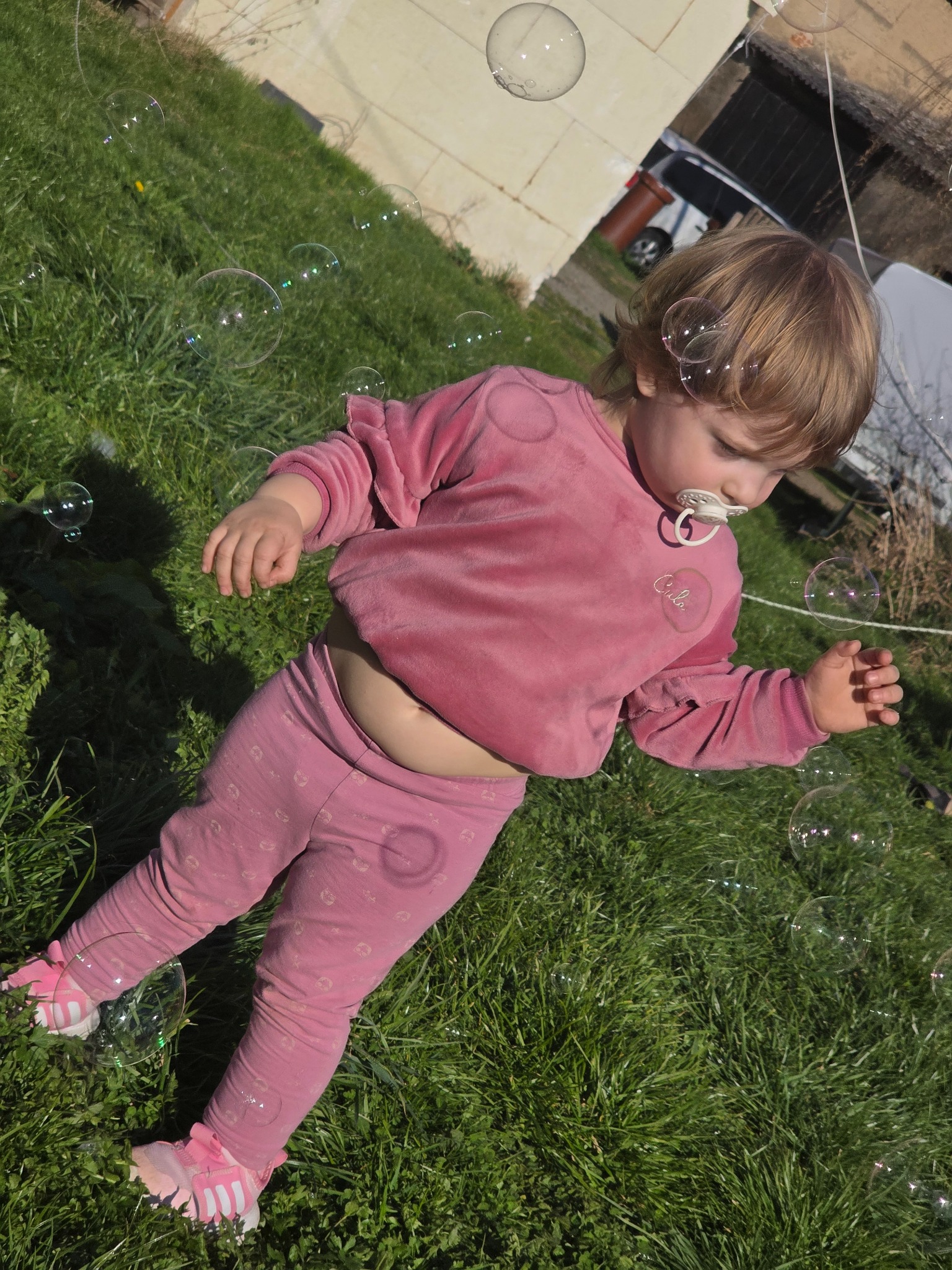 Mała dziewczynka w różowym ubranku bawi się na trawie z bańkami mydlanymi, w tle budynek.