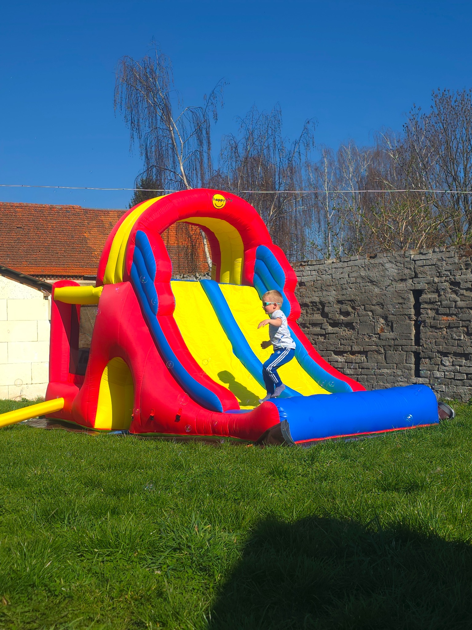 Chłopiec w okularach skacze na dmuchanej zjeżdżalni w kolorach czerwonym, żółtym i niebieskim, na trawniku przed ceglanym murem i budynkiem z czerwonym dachem.