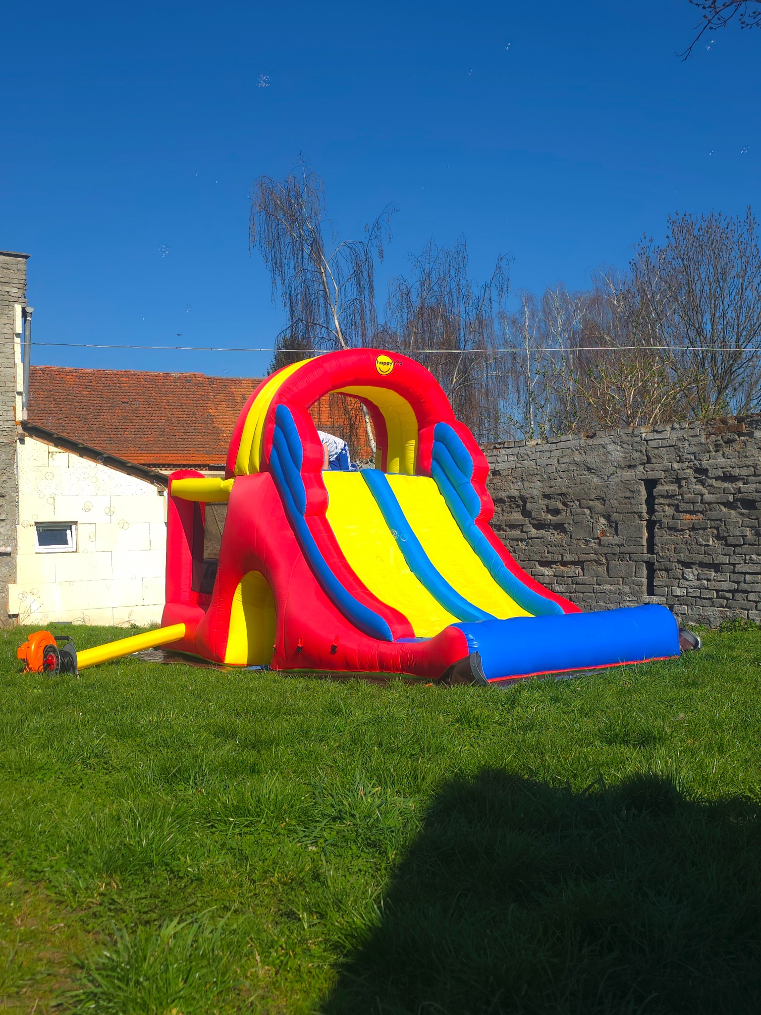 Rozłożona dmuchana zjeżdżalnia w kolorach czerwonym, żółtym i niebieskim na trawie, z podłączoną pompką elektryczną, na tle muru z kamienia i budynku z czerwoną dachówką.