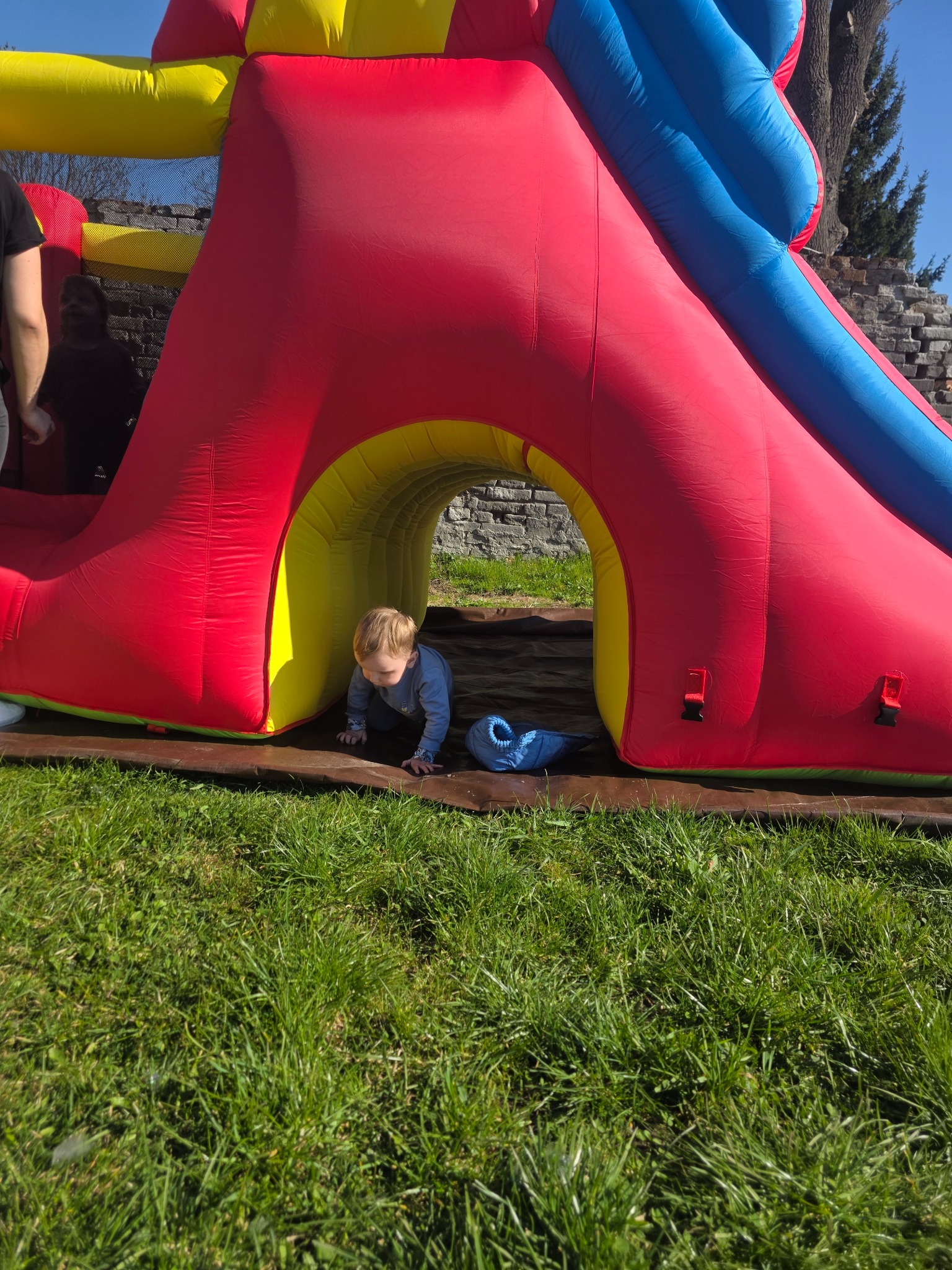 Chłopiec raczkuje w tunelu dmuchanego zamku z czerwoną ścianą, żółtym tunelem i niebieską zjeżdżalnią, na trawie w słoneczny dzień.