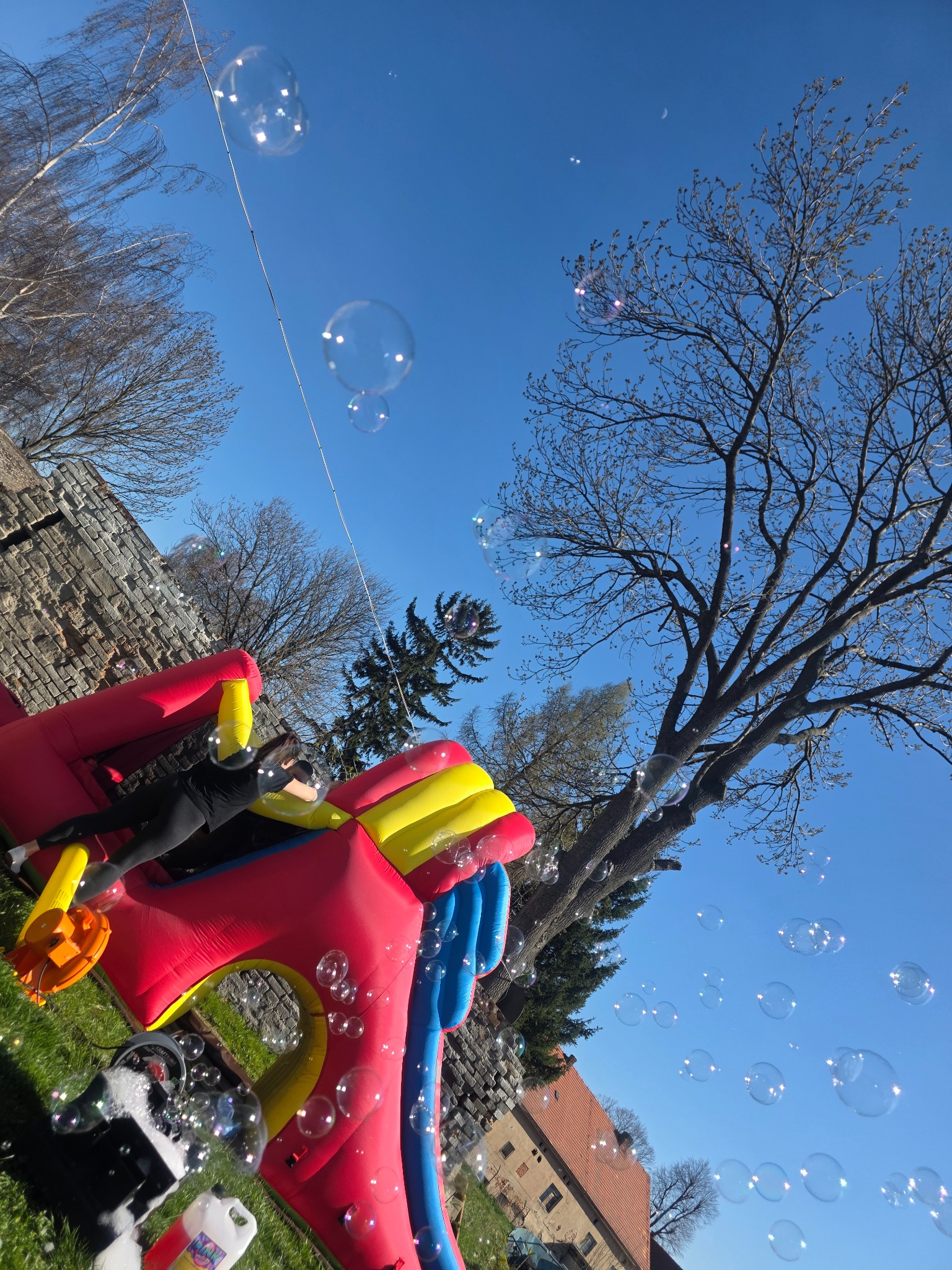 Dziewczynka w czarnym ubraniu leży na dmuchanym zamku w słoneczny dzień, w tle drzewa i budynki, a w powietrzu unoszą się bańki mydlane.