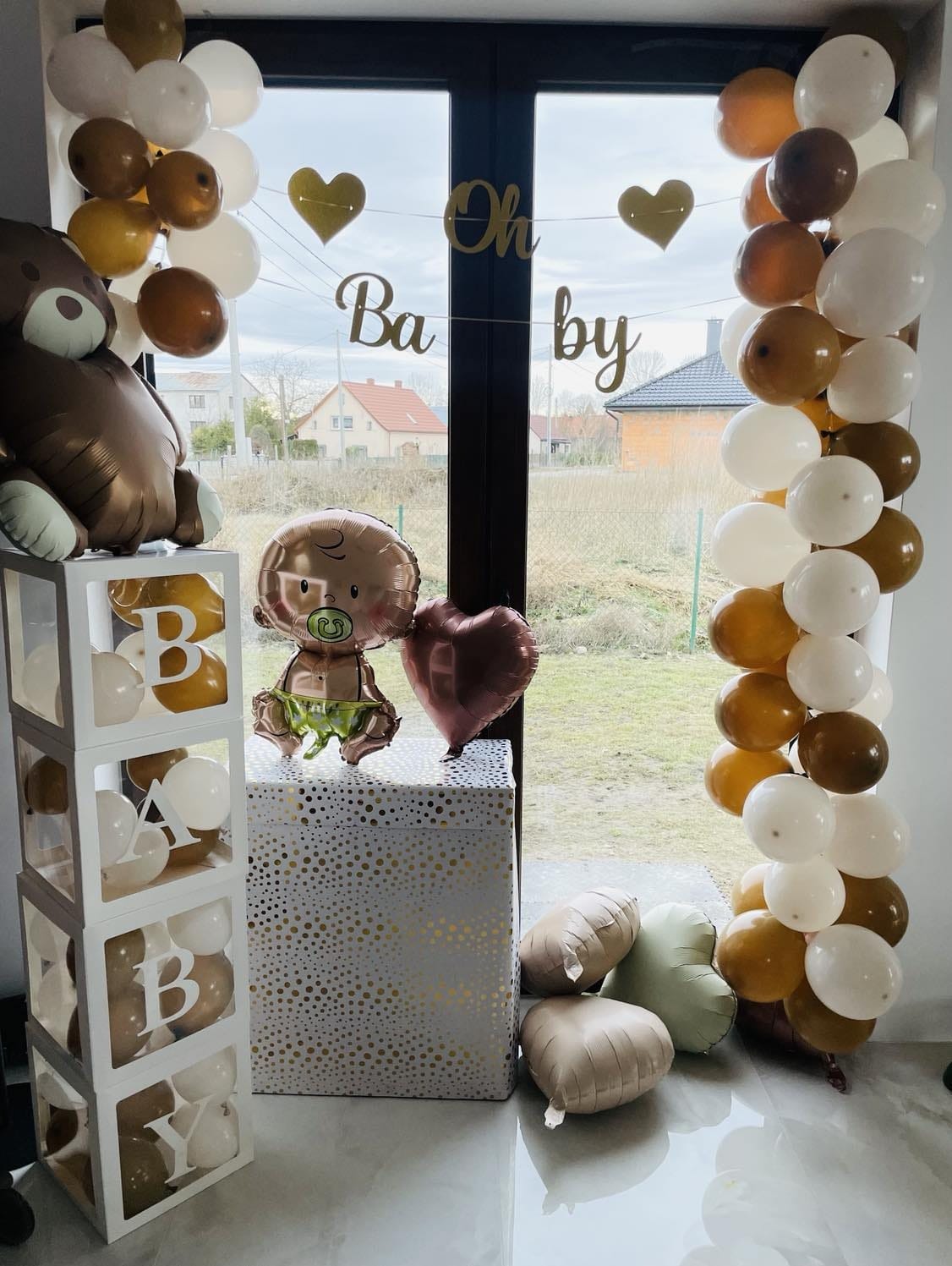 Dekoracja balonowa na przyjęcie baby shower w odcieniach brązu, bieli i złota, z balonami w kształcie misia, bobasa i serca, oraz napisem 'Oh Baby' na tle okna.