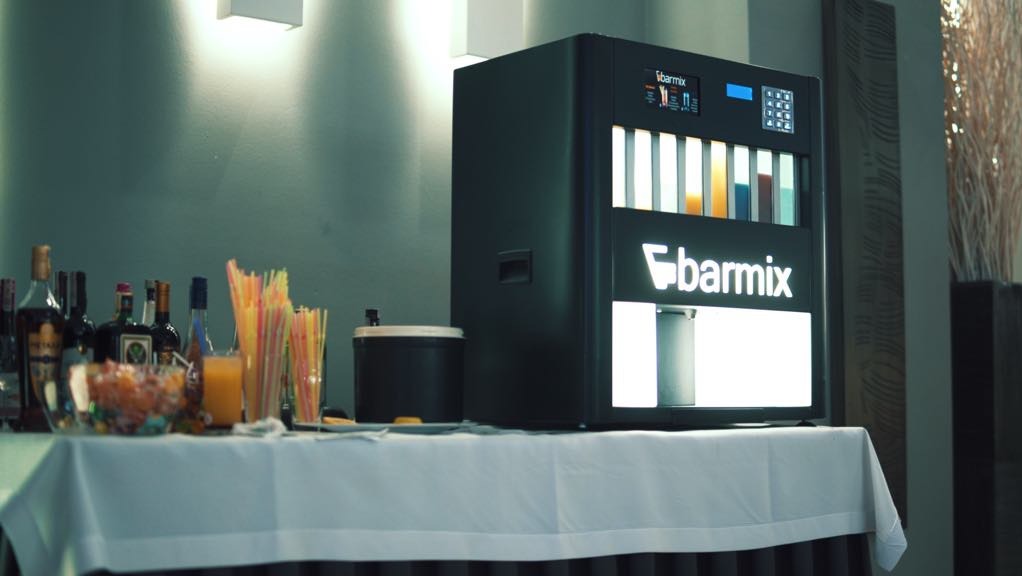 Automat do drinków Barmix na stole z alkoholem i słomkami, ustawiony podczas imprezy, widoczne podświetlane logo i kolorowe napoje w zbiornikach.