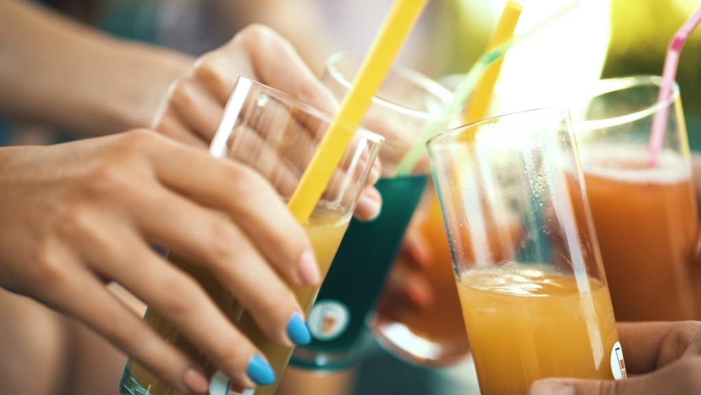 Zbliżenie na ręce trzymające szklanki z kolorowymi napojami i słomkami podczas toastu, w tle rozmyte światło słoneczne.