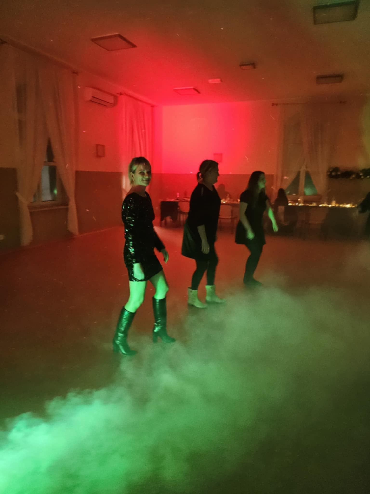 Kobieta w czarnej cekinowej sukience i wysokich butach tańczy na parkiecie w oparach zielonego dymu, w tle dwie inne kobiety i stoły z dekoracjami, oświetlenie czerwone i zielone.