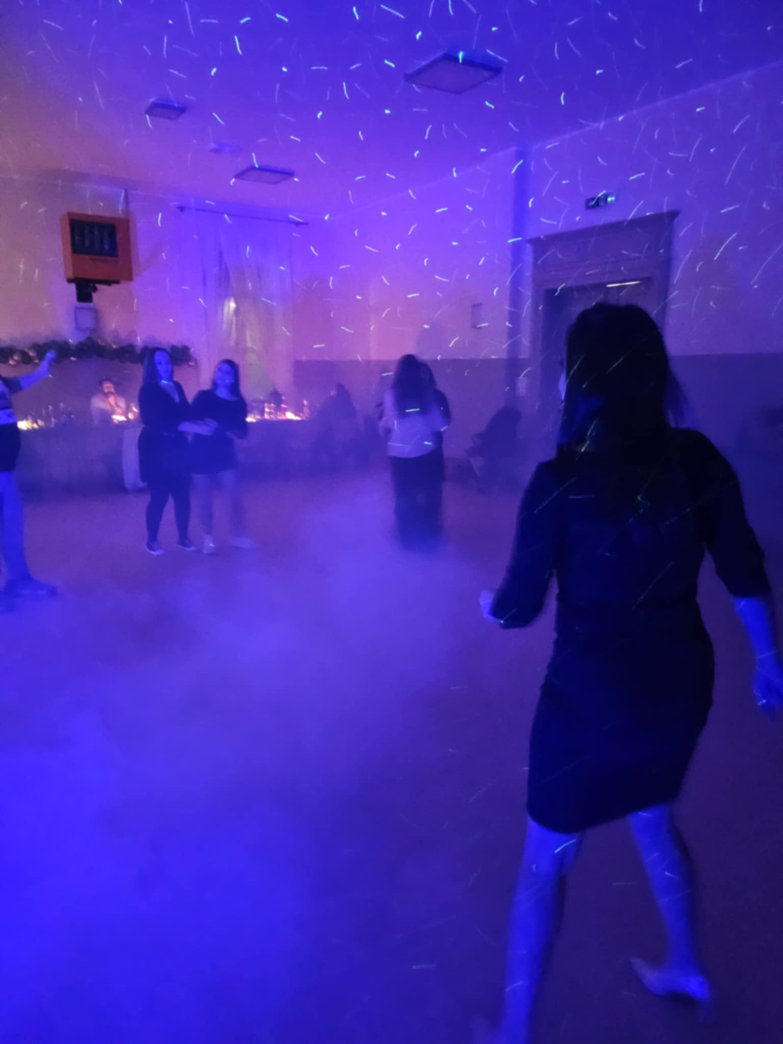 Wnętrze sali z efektem stroboskopowym, sylwetki osób w tle, pierwszoplanowa postać kobieca w ruchu, ciemne tło z punktowymi światłami.