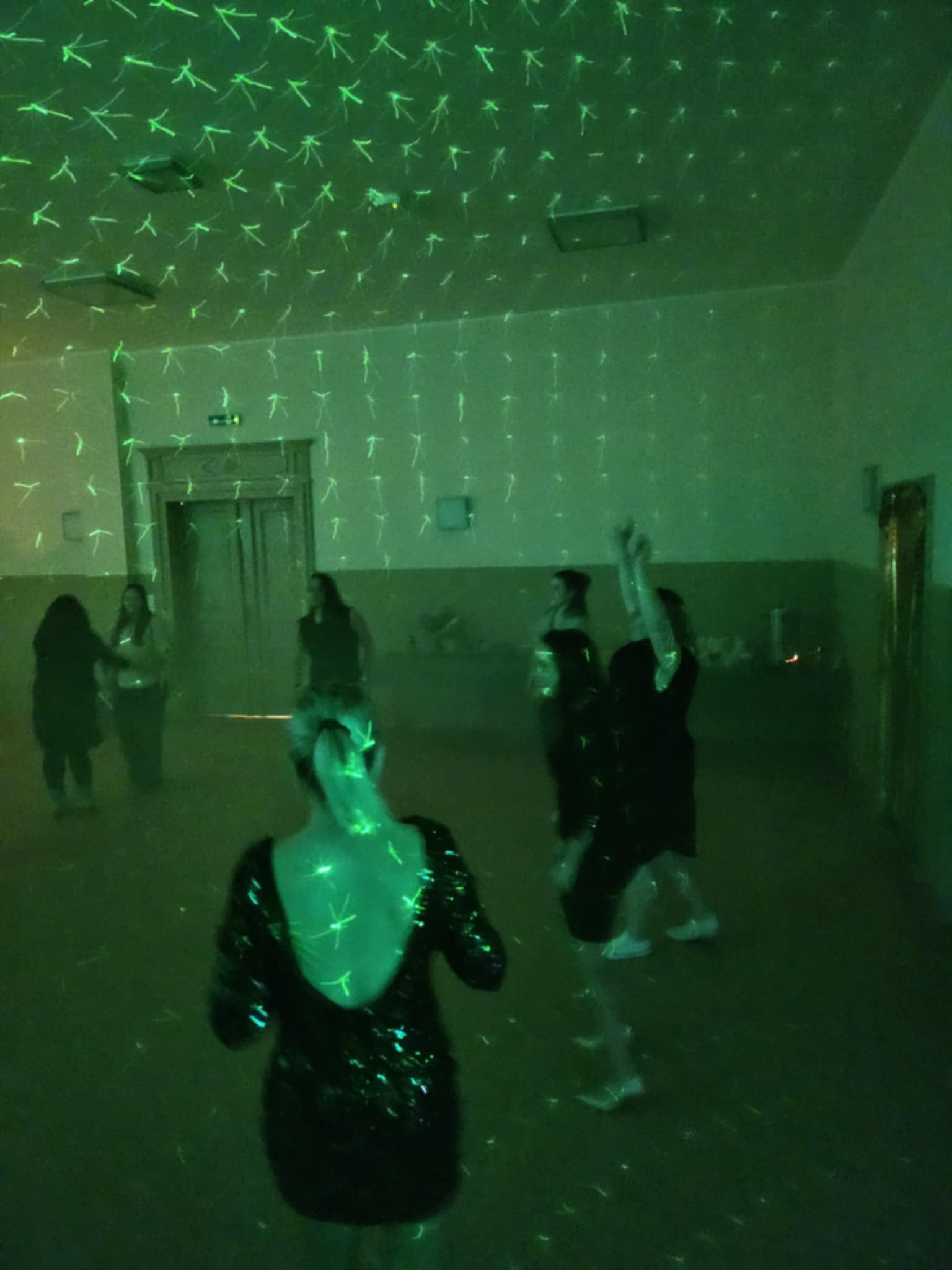Parkiet taneczny oświetlony zielonym laserem, grupa kobiet w tle, kobieta w cekinowej sukience z odkrytymi plecami na pierwszym planie.