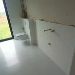 Wnęka z podwieszaną toaletą i heksagonalne płytki ścienne w łazience w trakcie wykańczania, widoczne złote baterie i niedokończone ściany.