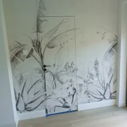 Ściana z fototapetą w motywy roślin tropikalnych, częściowo zasłonięta przez szklane drzwi zabezpieczone niebieską folią malarską.