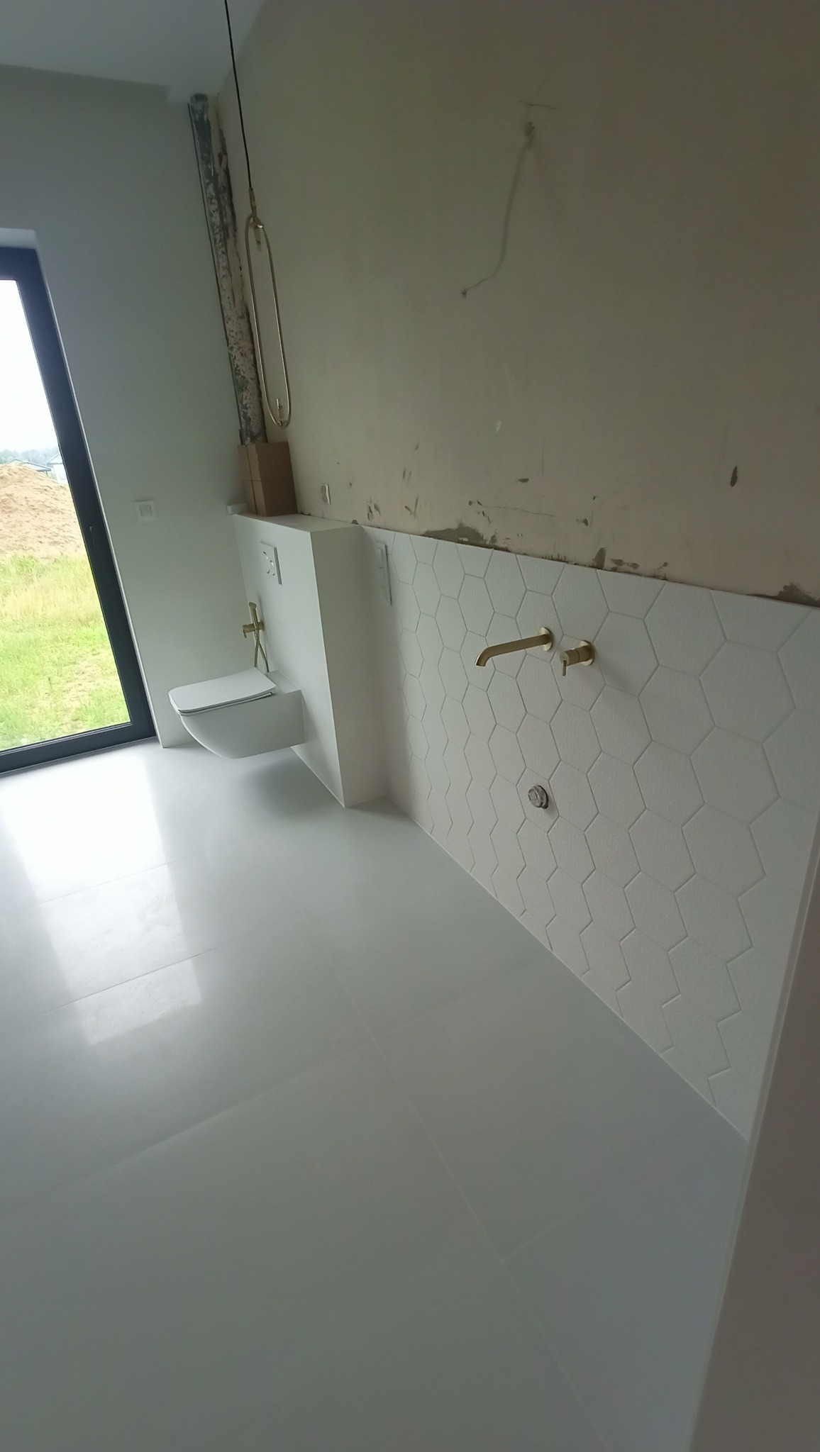 Wnęka z podwieszaną toaletą i heksagonalne płytki ścienne w łazience w trakcie wykańczania, widoczne złote baterie i niedokończone ściany.