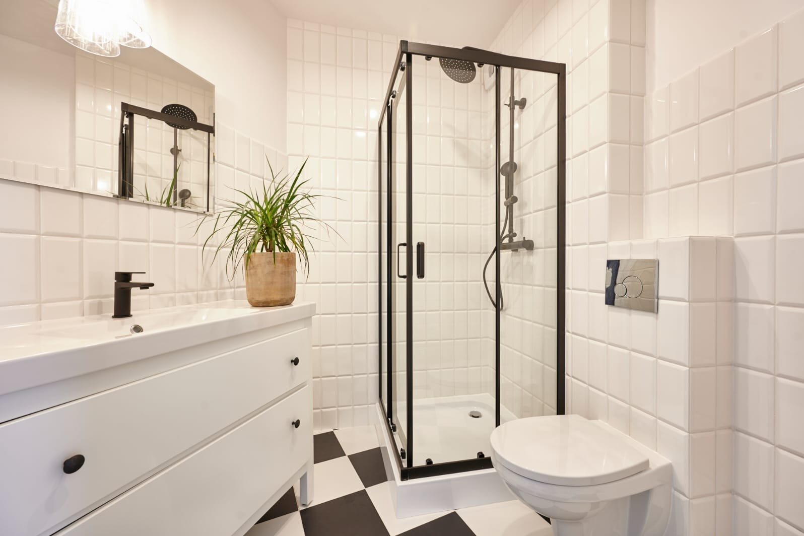 Nowoczesna łazienka z kabiną prysznicową w czarnej ramie, białą umywalką z czarną baterią, toaletą i podłogą w szachownicę, oświetlona lampą wiszącą.