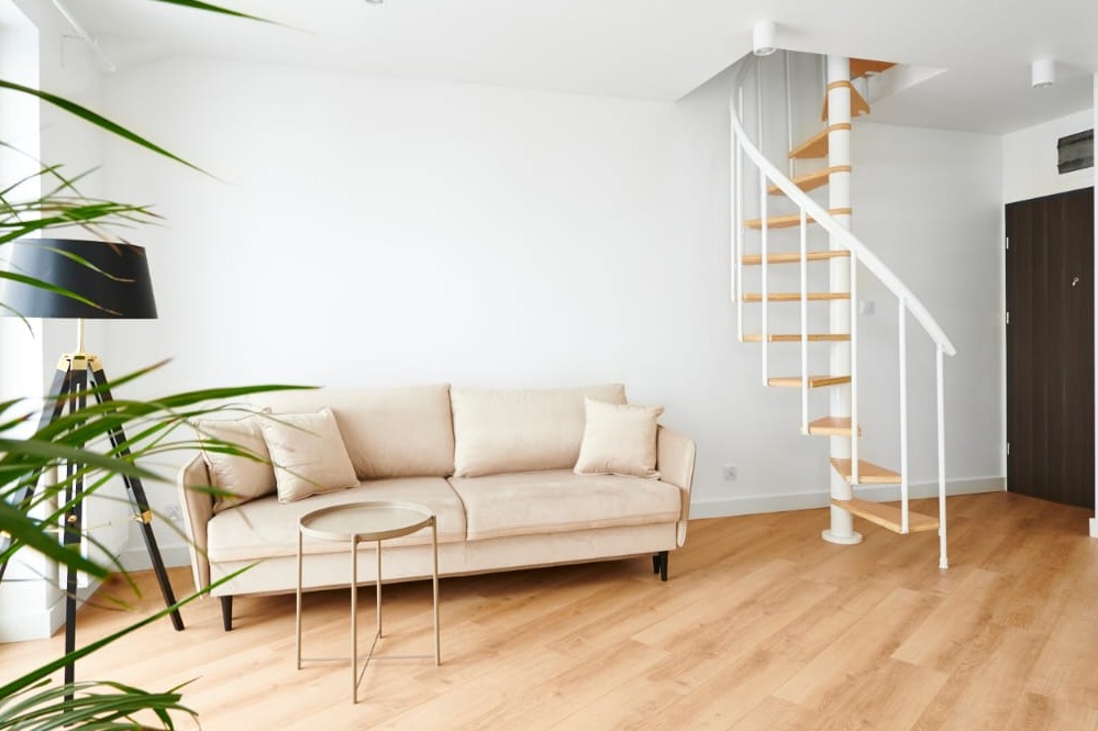 Jasny salon z drewnianą podłogą, beżową sofą, stolikiem kawowym i spiralnymi schodami na antresolę z drewnianymi stopniami i białymi barierkami.