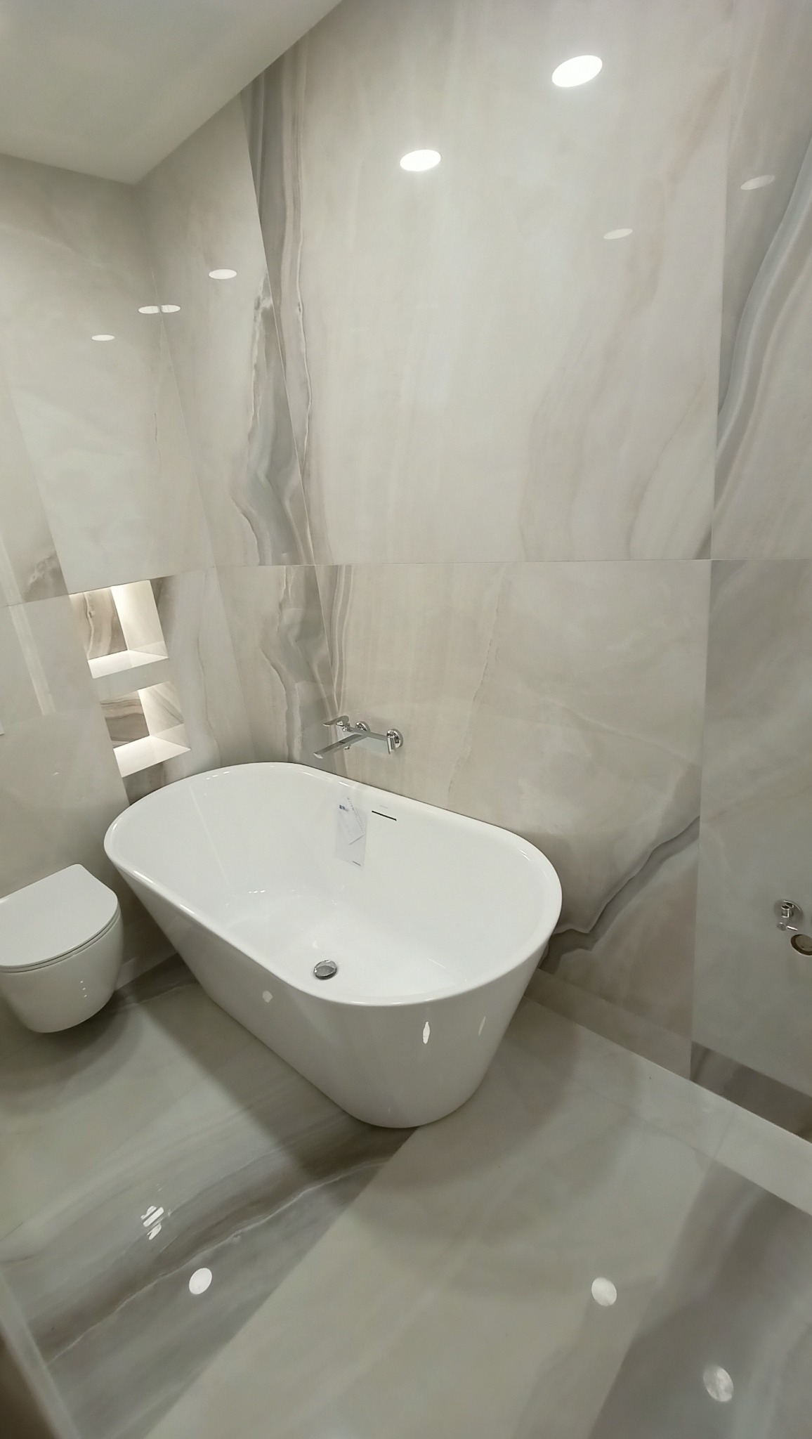 Nowoczesna, minimalistyczna łazienka z wolnostojącą białą wanną, podwieszaną toaletą i marmurowymi płytami w odcieniach szarości z subtelnym żyłkowaniem, oświetlona punktowymi lampami sufitowymi...