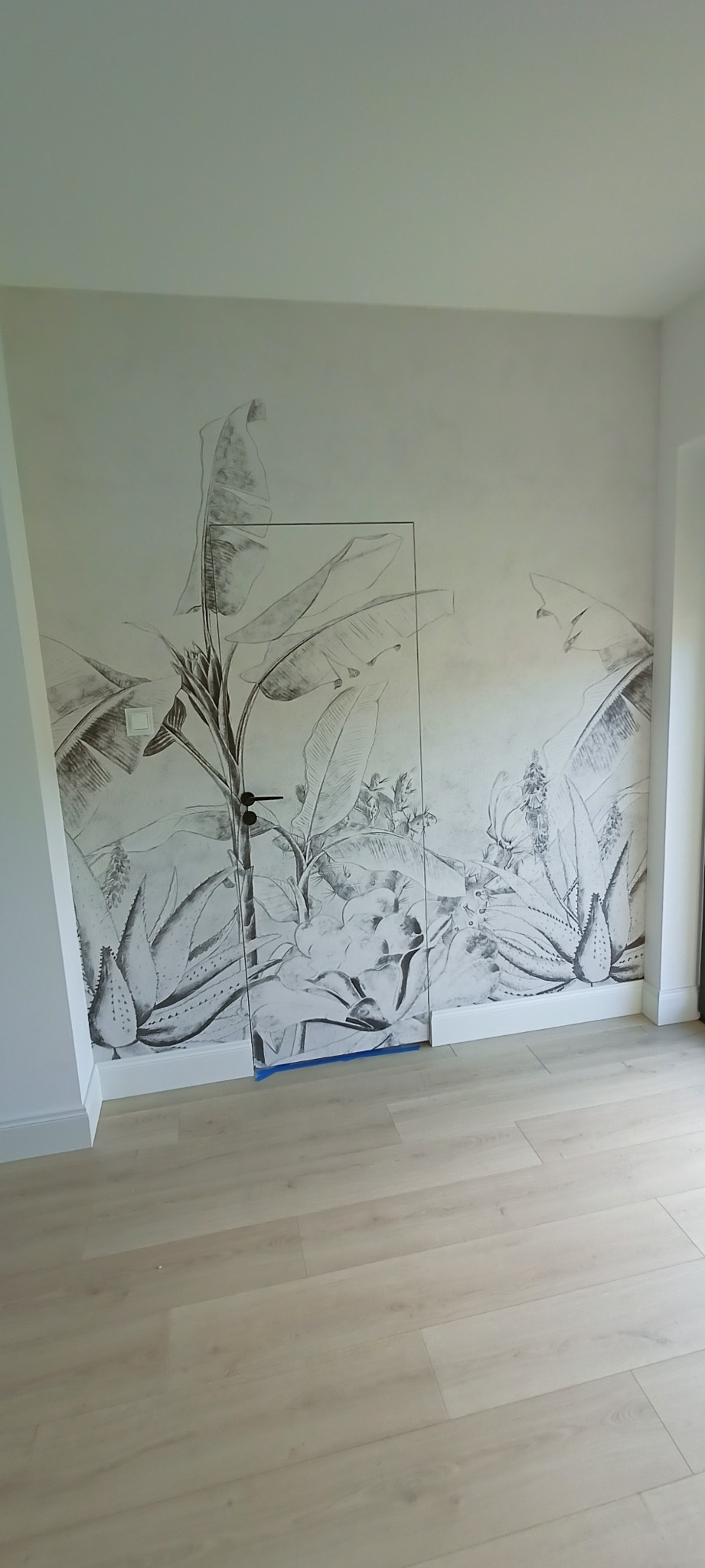 Ściana z fototapetą w motywy roślin tropikalnych, częściowo zasłonięta przez szklane drzwi zabezpieczone niebieską folią malarską.