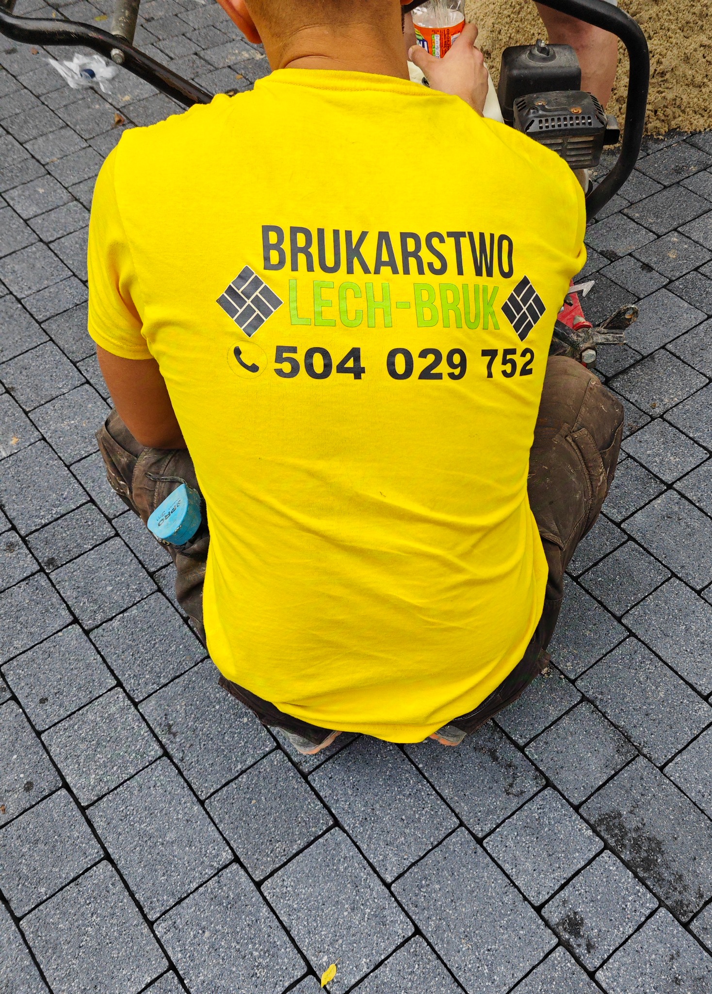 Brukarz w żółtej koszulce z logo firmy, klęczy na ułożonej szarej kostce brukowej, obok zagęszczarka wibracyjna i miarka w kieszeni spodni.