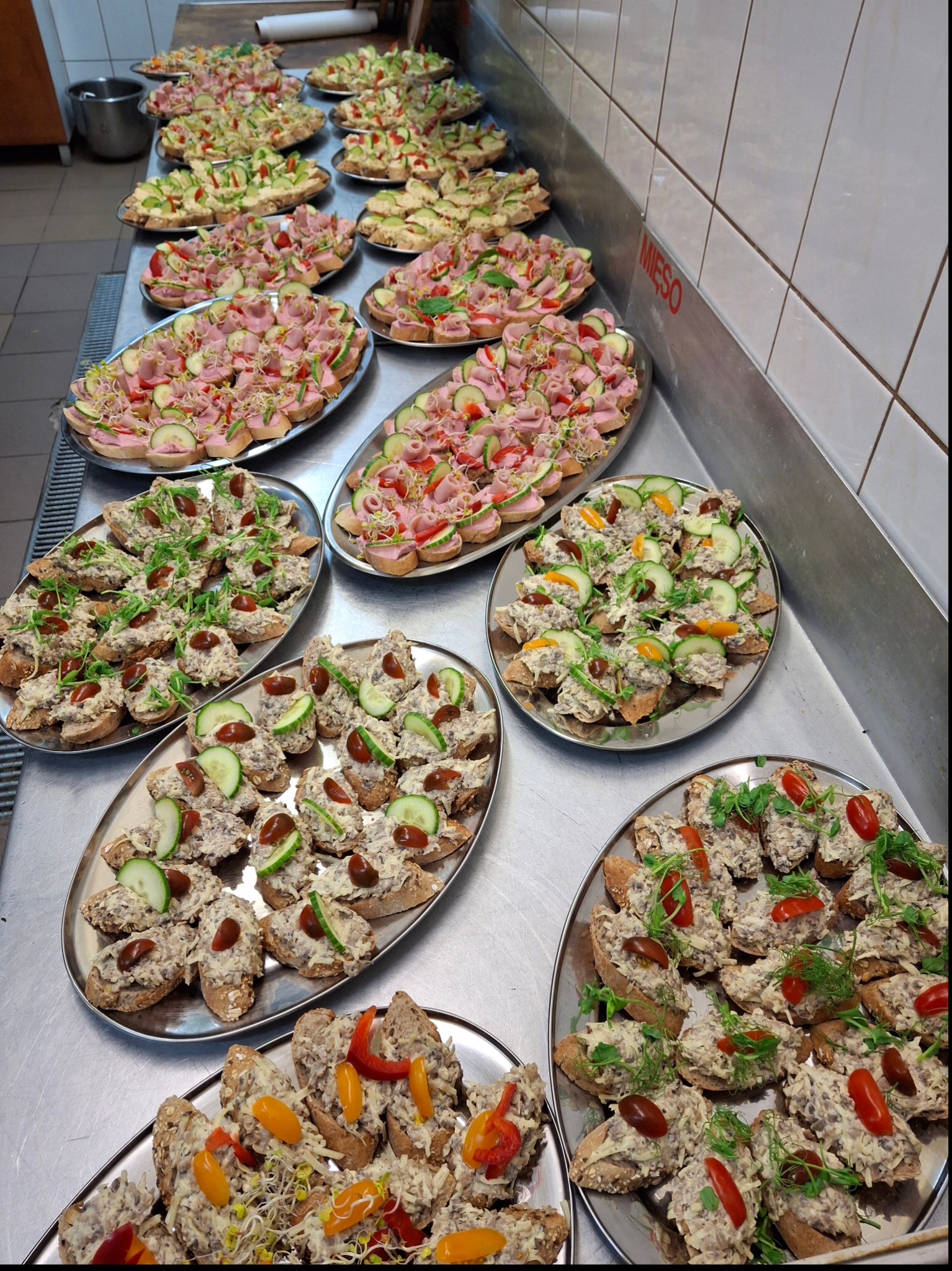 Rzędy srebrnych tac z różnorodnymi kanapkami na kromkach chleba, udekorowanymi warzywami i kiełkami, ustawione na metalowym stole w zapleczu kuchennym z białymi kafelkami.