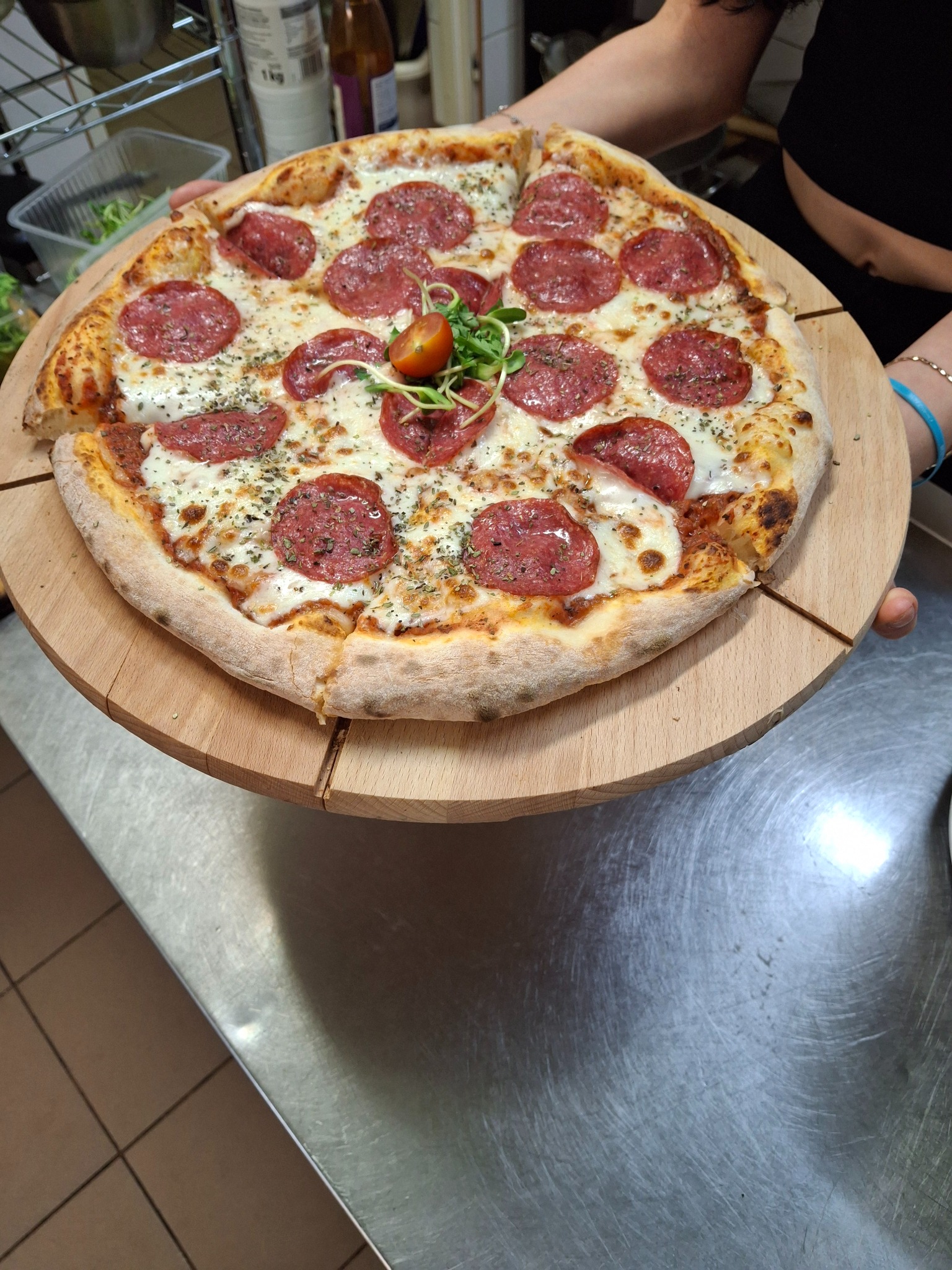 Pizza pepperoni z mozarellą, posypana ziołami i udekorowana kiełkami oraz połówką pomidora, serwowana na drewnianej desce.