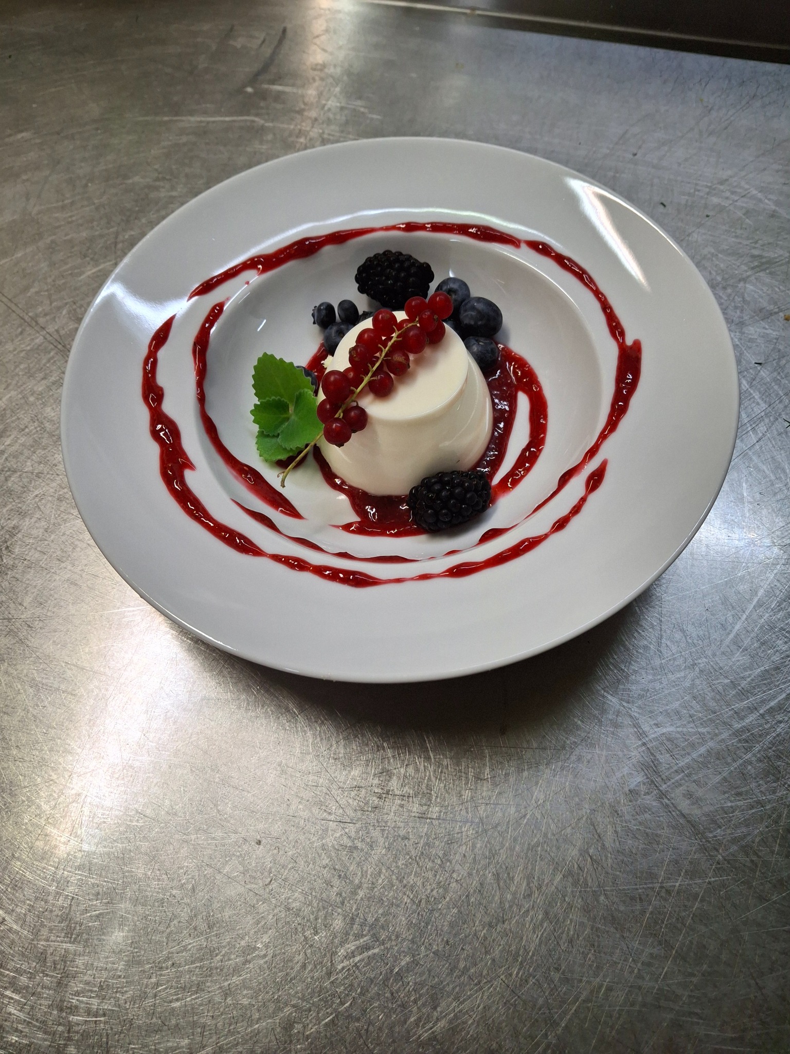 Elegancko podana panna cotta na białym talerzu, udekorowana sosem owocowym, jeżynami, borówkami, czerwoną porzeczką i listkiem mięty, na tle metalowego blatu.