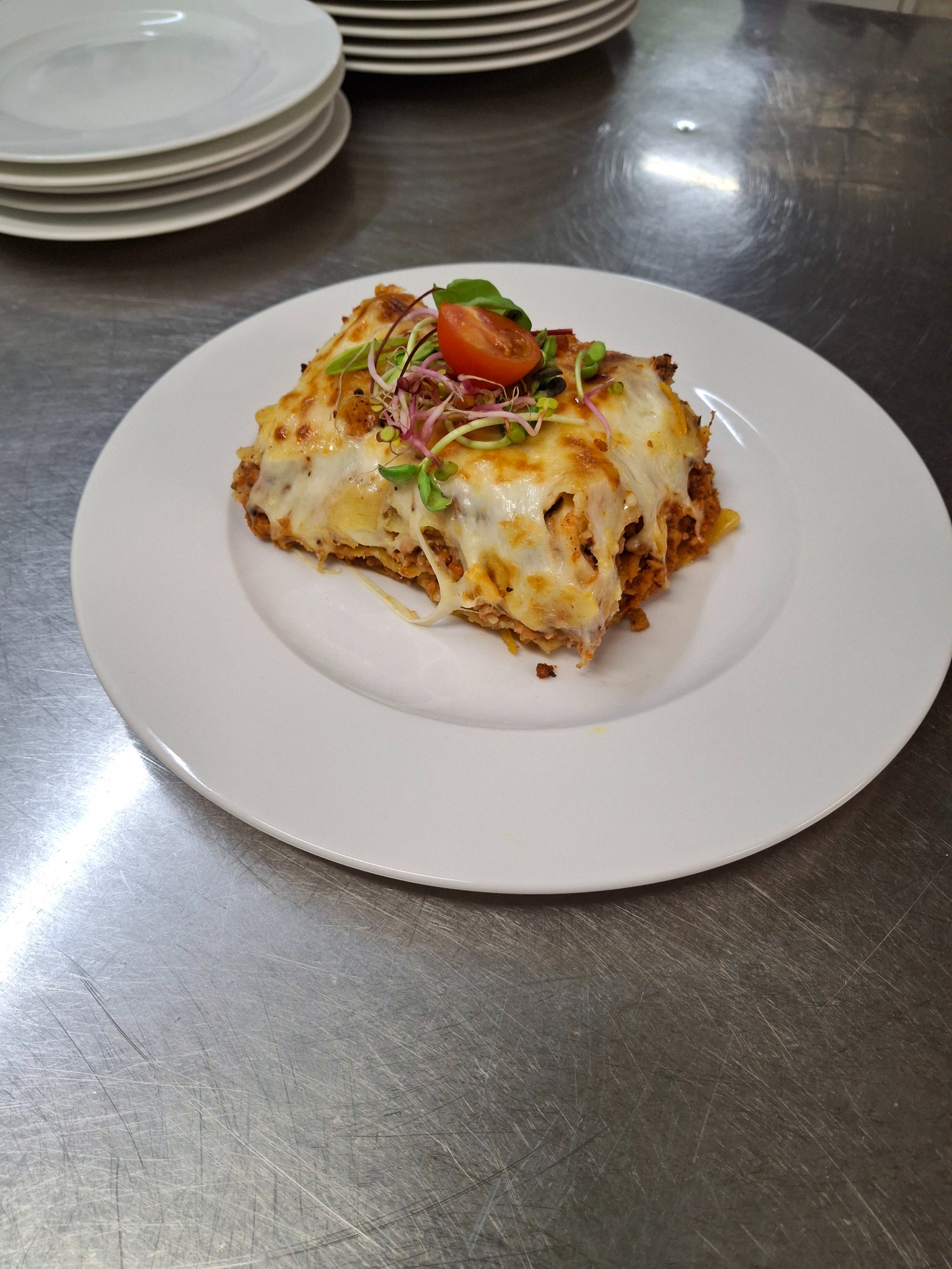 Porcja lasagne na białym talerzu, udekorowana kiełkami i połówką pomidora koktajlowego, na tle metalowego blatu roboczego i stosu talerzy.